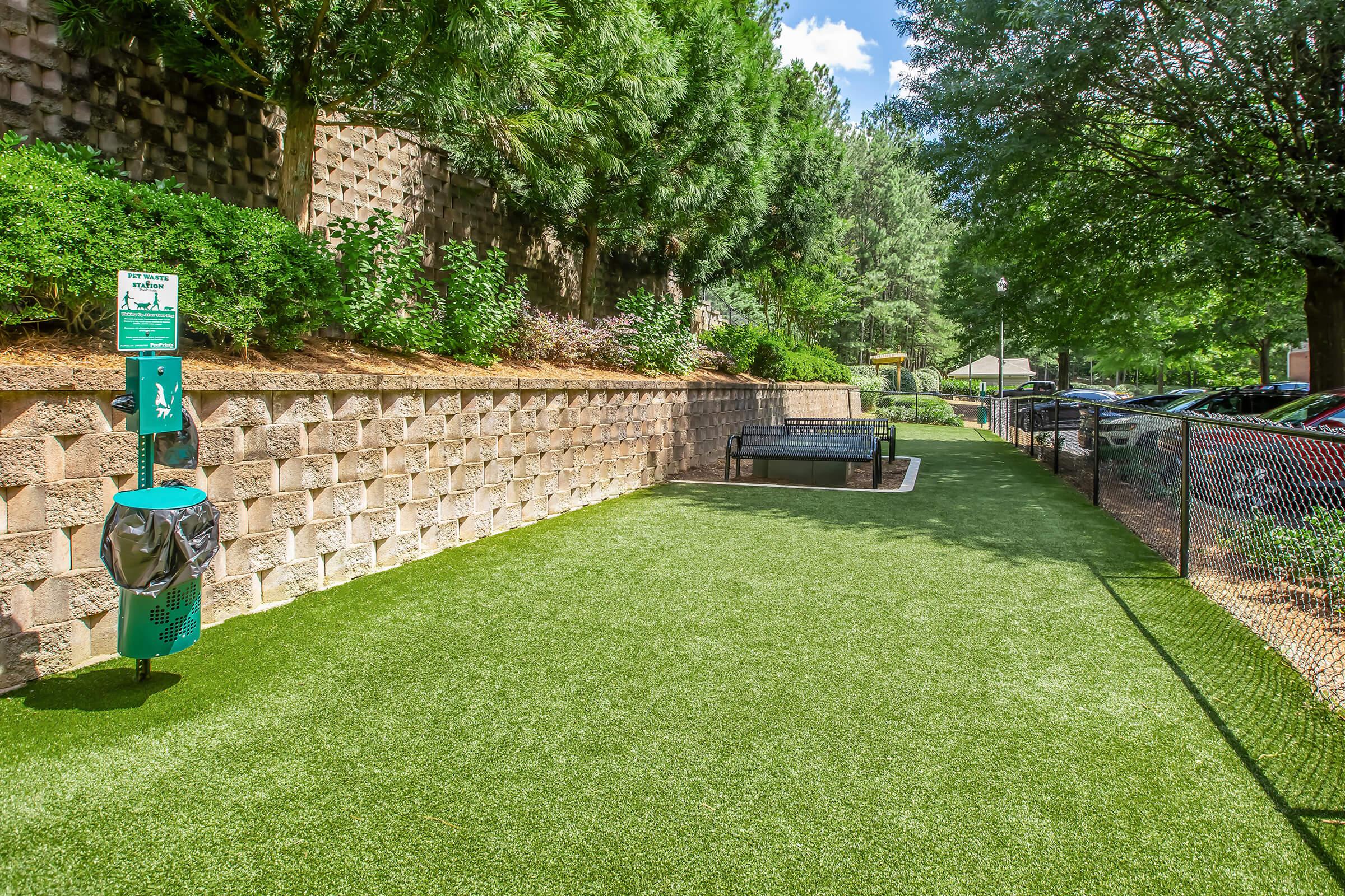 A well-maintained grassy area with artificial turf, featuring a green pet waste station, a trash bin, and a bench surrounded by trees and shrubs. In the background, a fence borders the section, and there's a glimpse of parked cars and a pathway. The scene is under a clear blue sky with a few clouds.