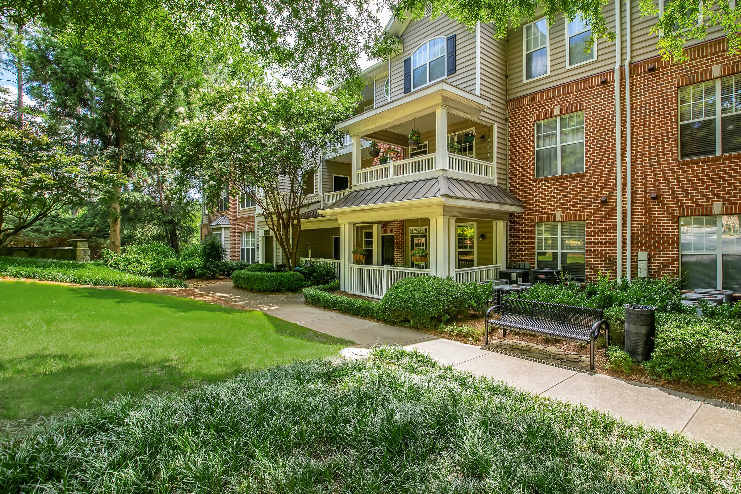 A peaceful apartment complex exterior featuring a brick facade, multiple balconies, and surrounded by lush greenery. There is a well-maintained lawn, flowering plants, a walkway, and a bench along the path, creating a welcoming atmosphere for residents and visitors.