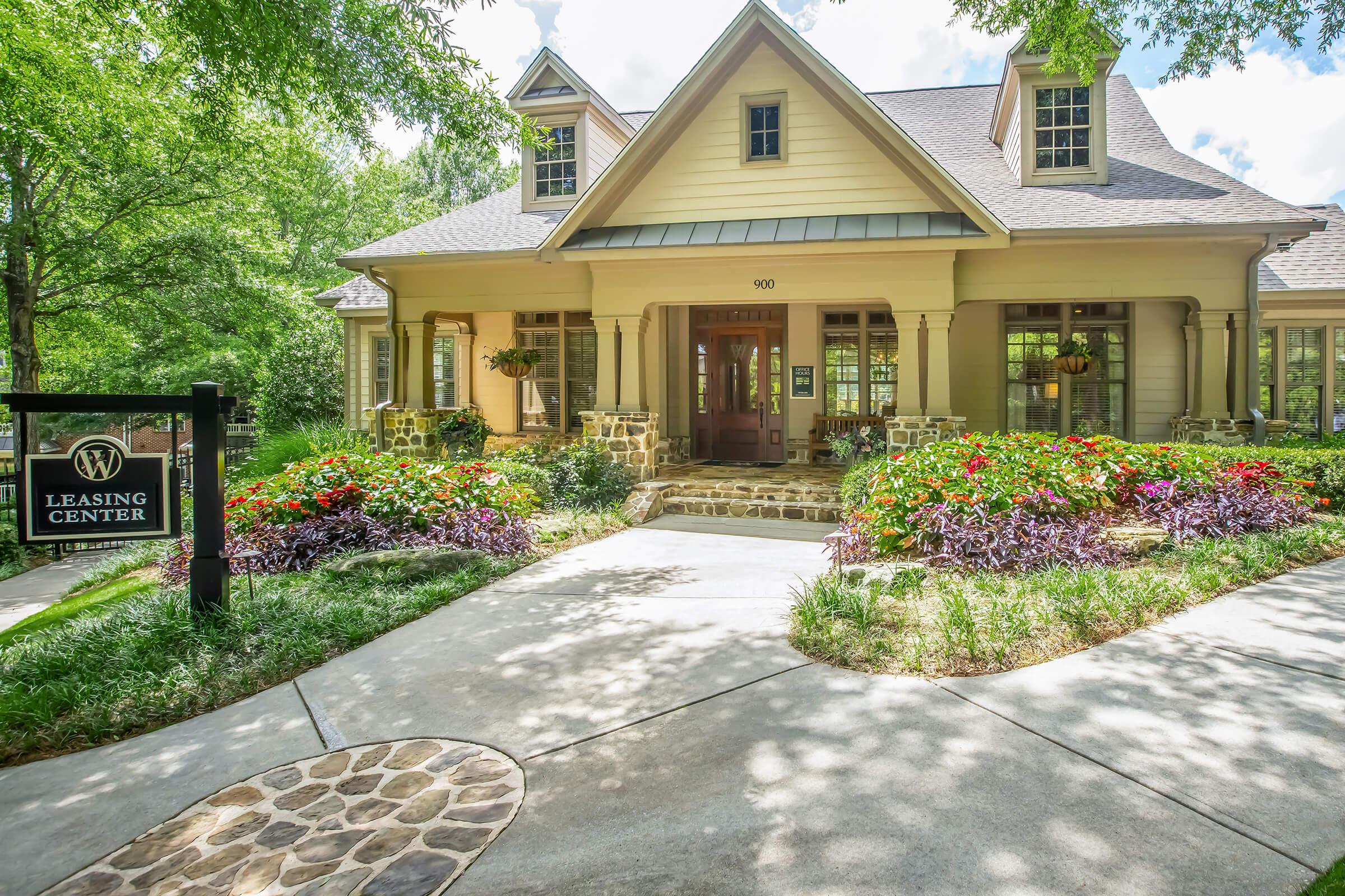 A charming leasing center with a welcoming entrance surrounded by vibrant flower beds and lush greenery. The building features a stylish design with large windows, a peaked roof, and a cozy porch. A sign in front clearly indicates "Leasing Center," inviting visitors to explore the property.