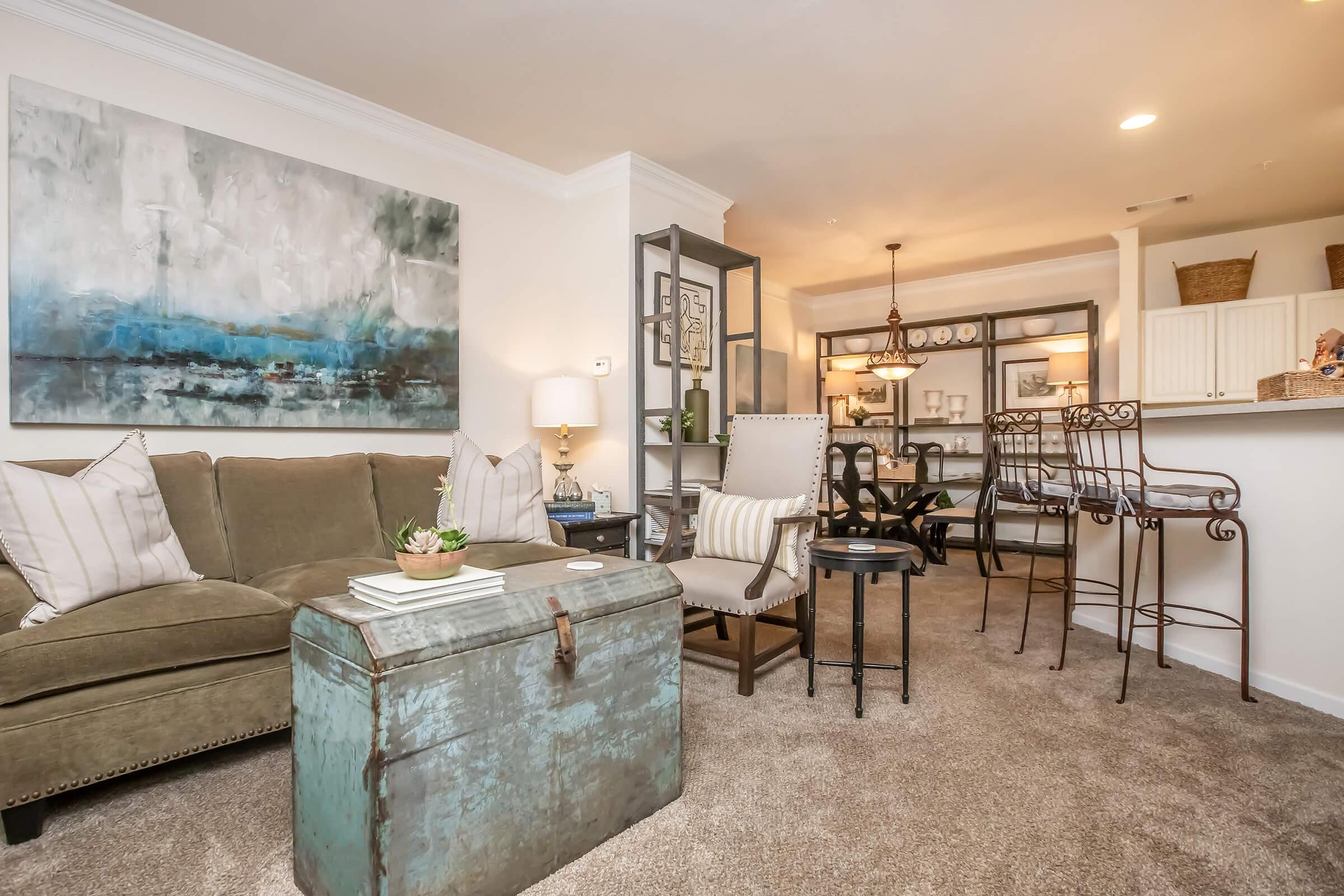Cozy living room featuring a green sofa with decorative pillows, a vintage blue chest as a coffee table, and a beige armchair. A textured painting hangs on the wall, while a dining area with a table and chairs is visible in the background, complemented by warm lighting and stylish decor.