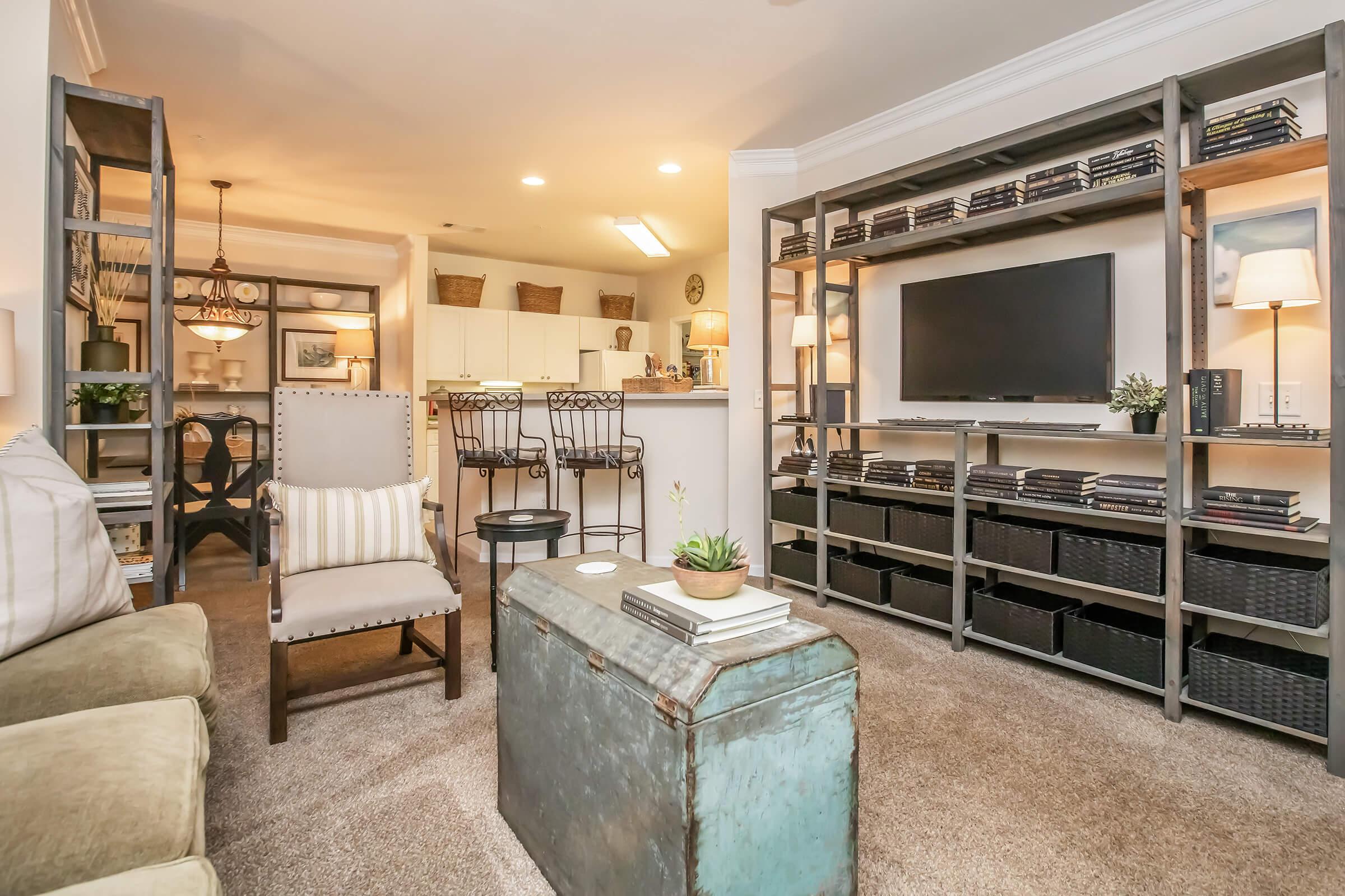 Cozy living room featuring a green armchair, a coffee table made from an antique trunk, and a modern entertainment center. The space is well-lit with lamps, and there's a dining area visible in the background. Shelves hold books and decorative items, adding a personal touch to the decor.