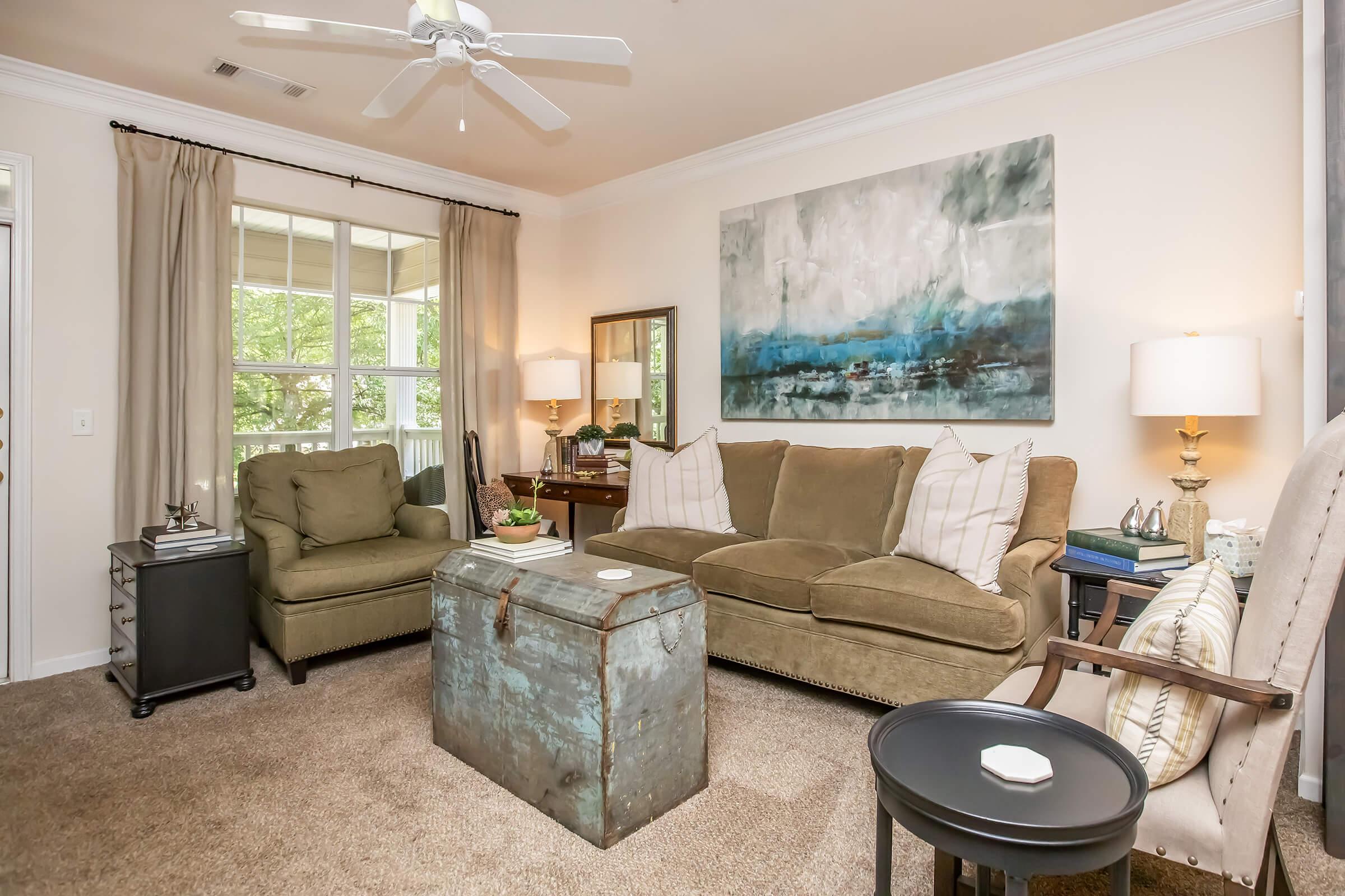 Cozy living room featuring a green sofa with decorative pillows, a vintage trunk as a coffee table, and a small black side table. Natural light streams through large windows with beige curtains. The walls are adorned with a large abstract painting, and there are two armchairs and a mirror enhancing the space.