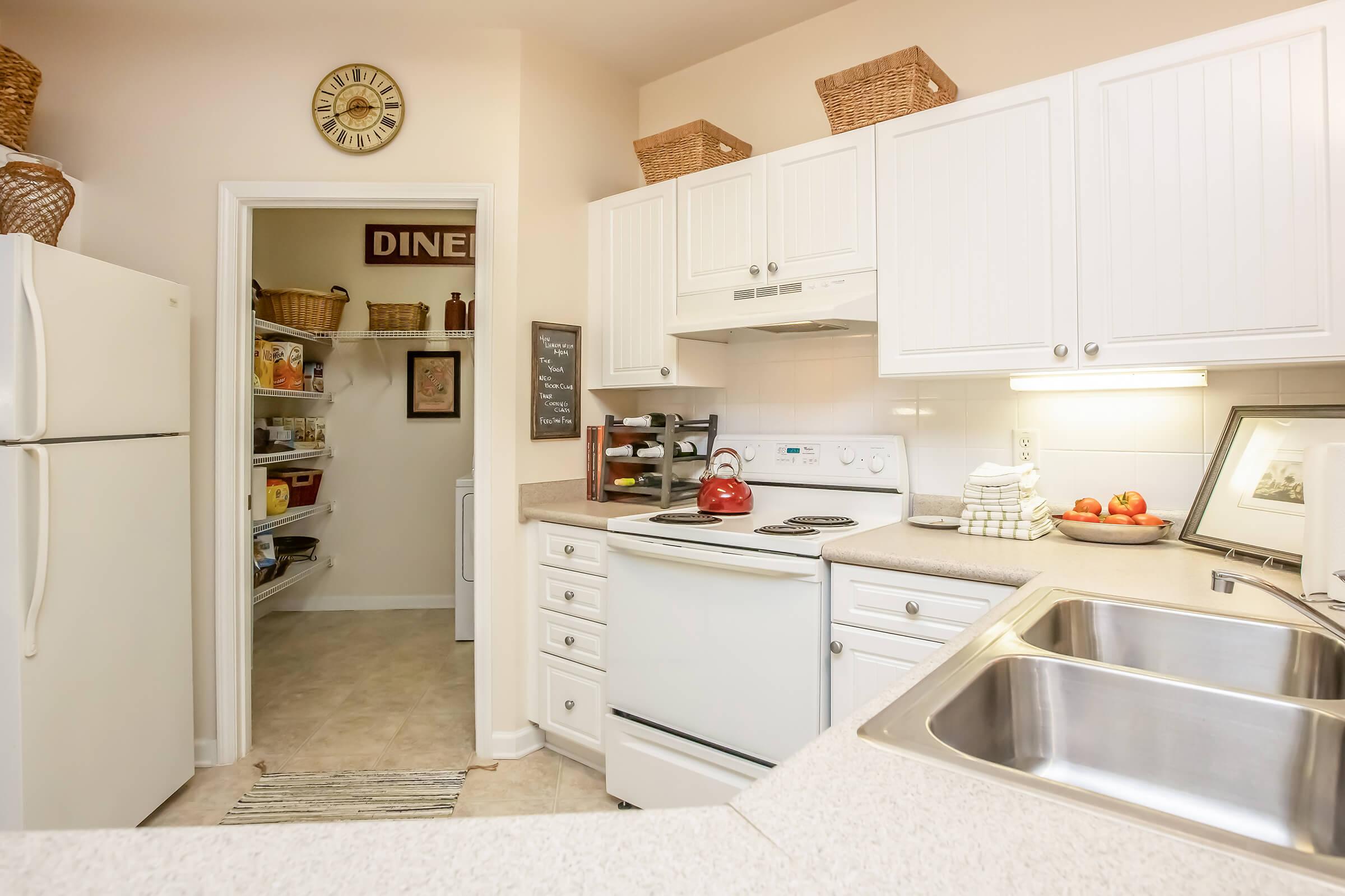 A bright, modern kitchen featuring white cabinetry, a stainless steel sink, and an oven. There is a red kettle on the stove, neatly arranged kitchen towels, and a bowl of apples on the counter. A pantry door is visible in the background, showcasing organized shelves. A clock and decorative baskets add a cozy touch.