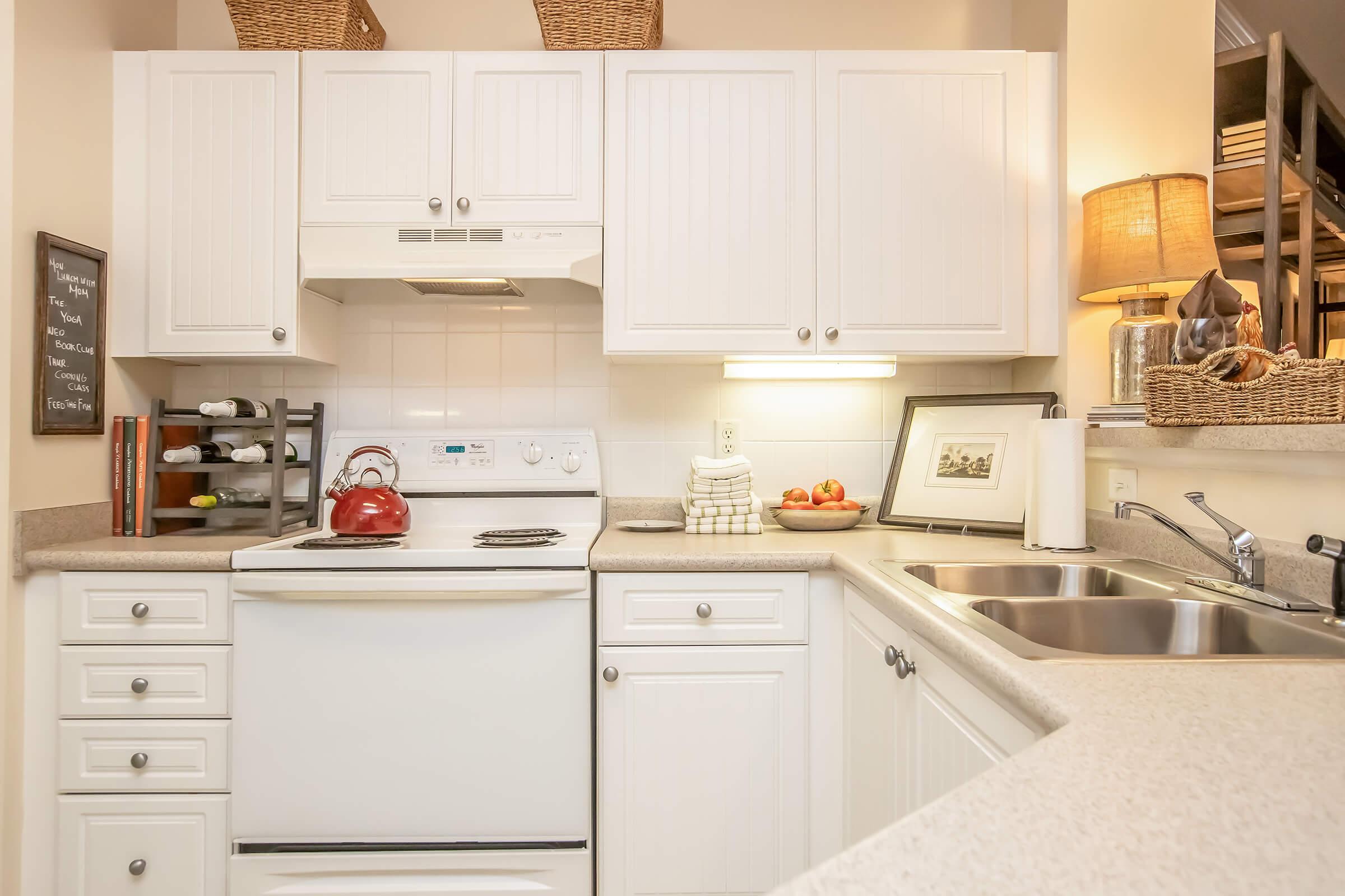 A cozy kitchen featuring white cabinetry, a red kettle on the stove, and a double sink. There are wine bottles in a rack, clean dish towels, and a bowl of apples on the countertop. A lamp provides warm lighting, and shelves hold decorative baskets, creating a welcoming atmosphere.