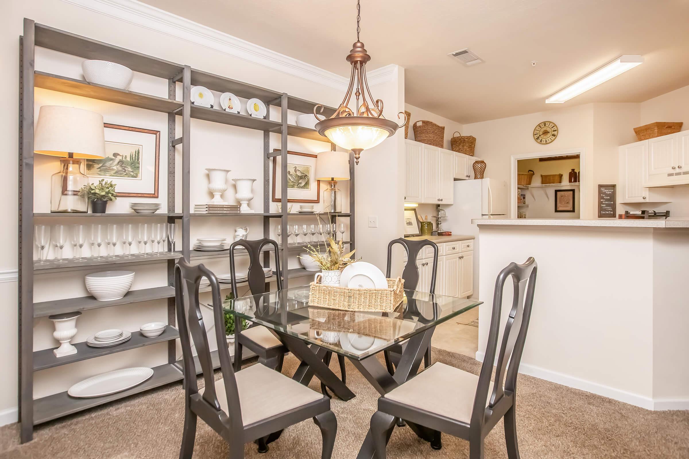 A well-decorated dining area featuring a glass table surrounded by four chairs. Behind the table, there is a stylish gray shelving unit displaying various decorative items and dishware. A warm chandelier hangs above, providing light to the space, which has a cozy, welcoming atmosphere. The kitchen is visible in the background.