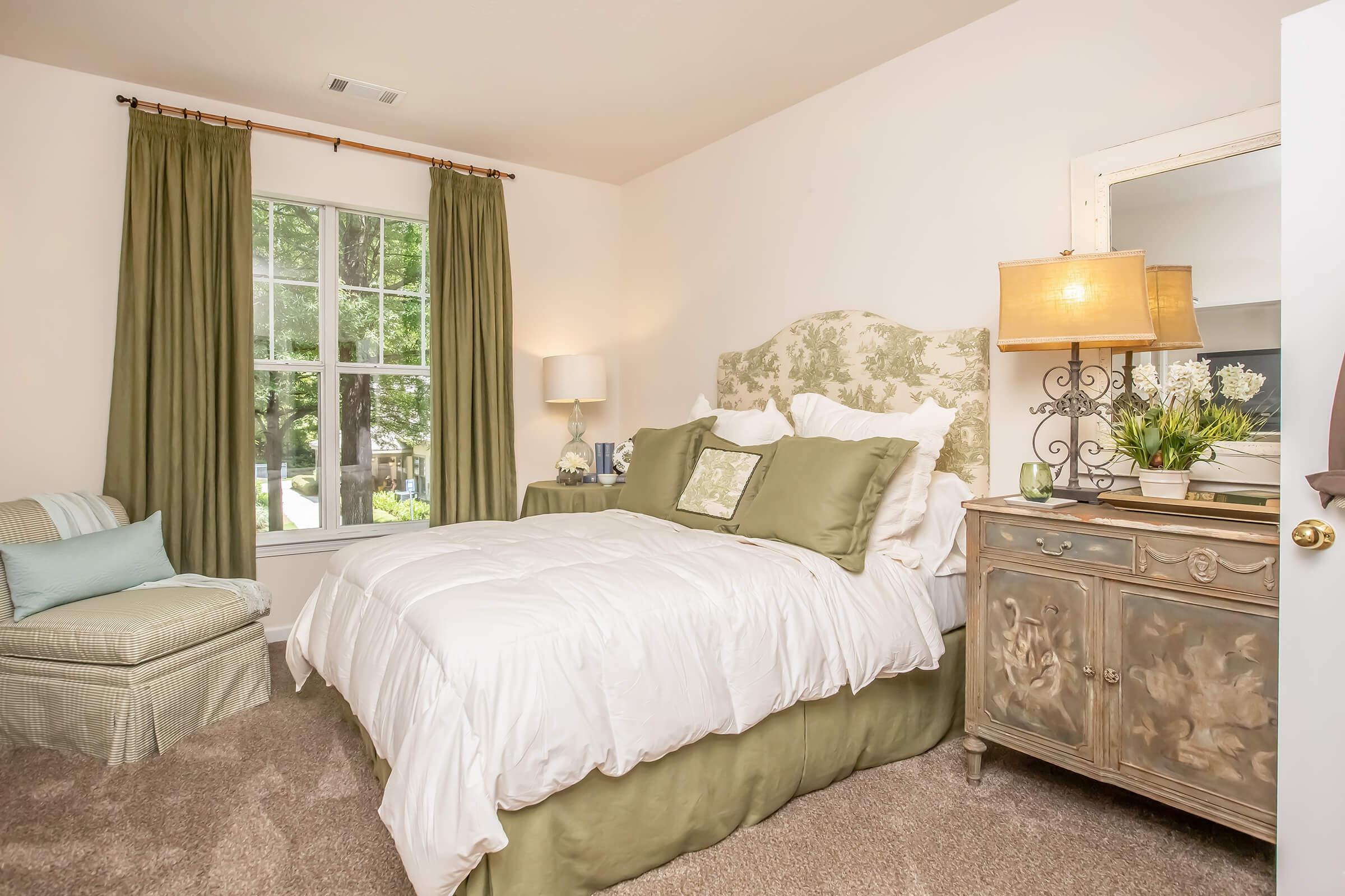 A cozy bedroom featuring a soft green and white color scheme. It has a plush bed with decorative pillows, a vintage-style dresser, and an accent chair. Large windows allow natural light, framed by green curtains. A bedside lamp adds warmth to the inviting atmosphere.