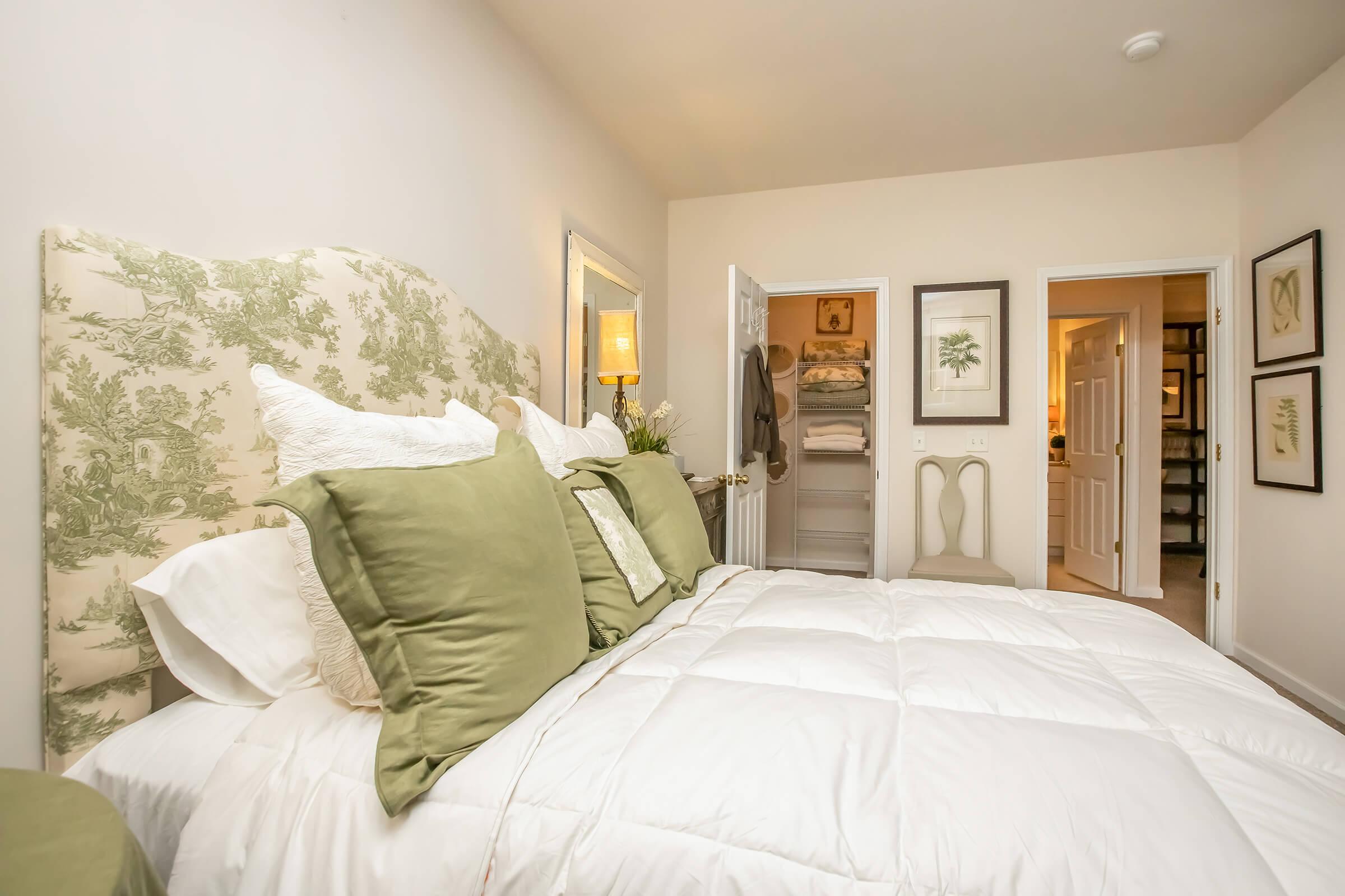 A cozy bedroom featuring a plush bed with a decorative green headboard and large green pillows. The bed is topped with a white comforter. In the background, there is a glimpse of a closet and a bathroom door, with framed artwork on the walls, creating a serene and inviting atmosphere.