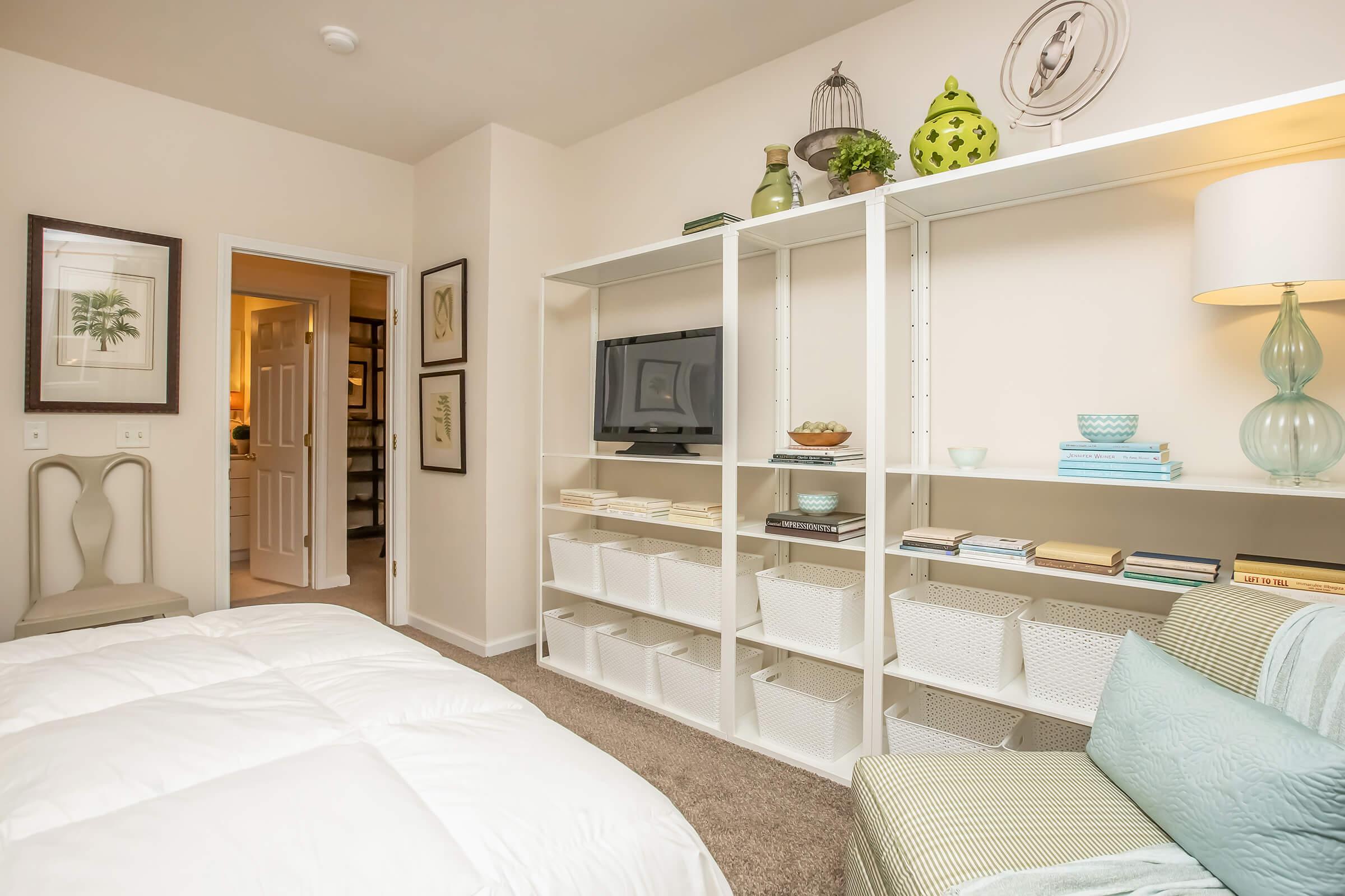 A cozy bedroom featuring a neatly made bed with white bedding, a light green accent chair, and a white shelving unit displaying decorative items and storage baskets. A TV is placed on the shelf, and framed art adorns the walls. A doorway leads to a bathroom area, adding to the room’s inviting atmosphere.