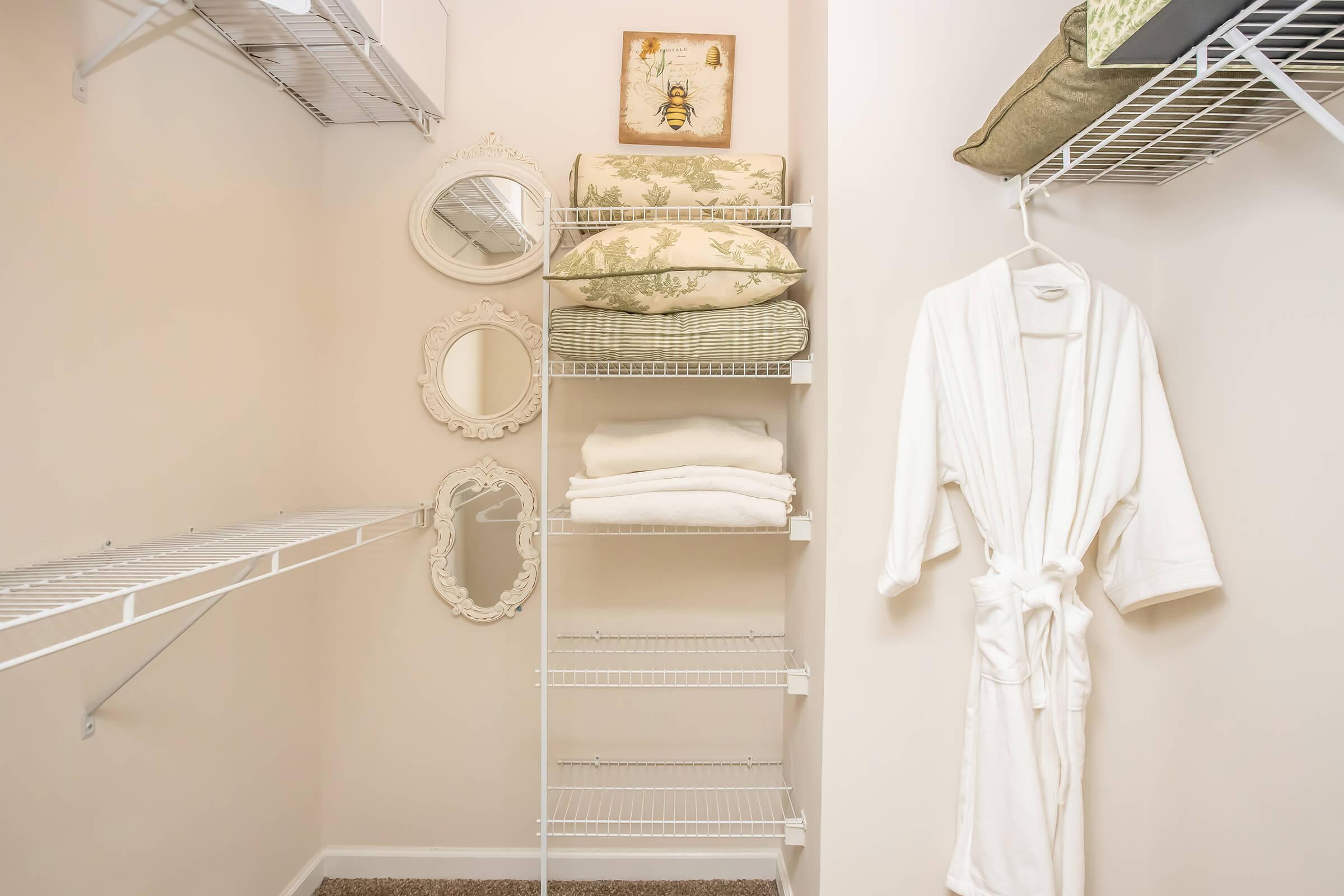A neatly organized closet featuring white wire shelving holding stacked pillows and towels. Two small decorative mirrors are mounted on the wall, and a white robe hangs on the right side. The walls are painted a soft neutral color, creating a clean and calming atmosphere.