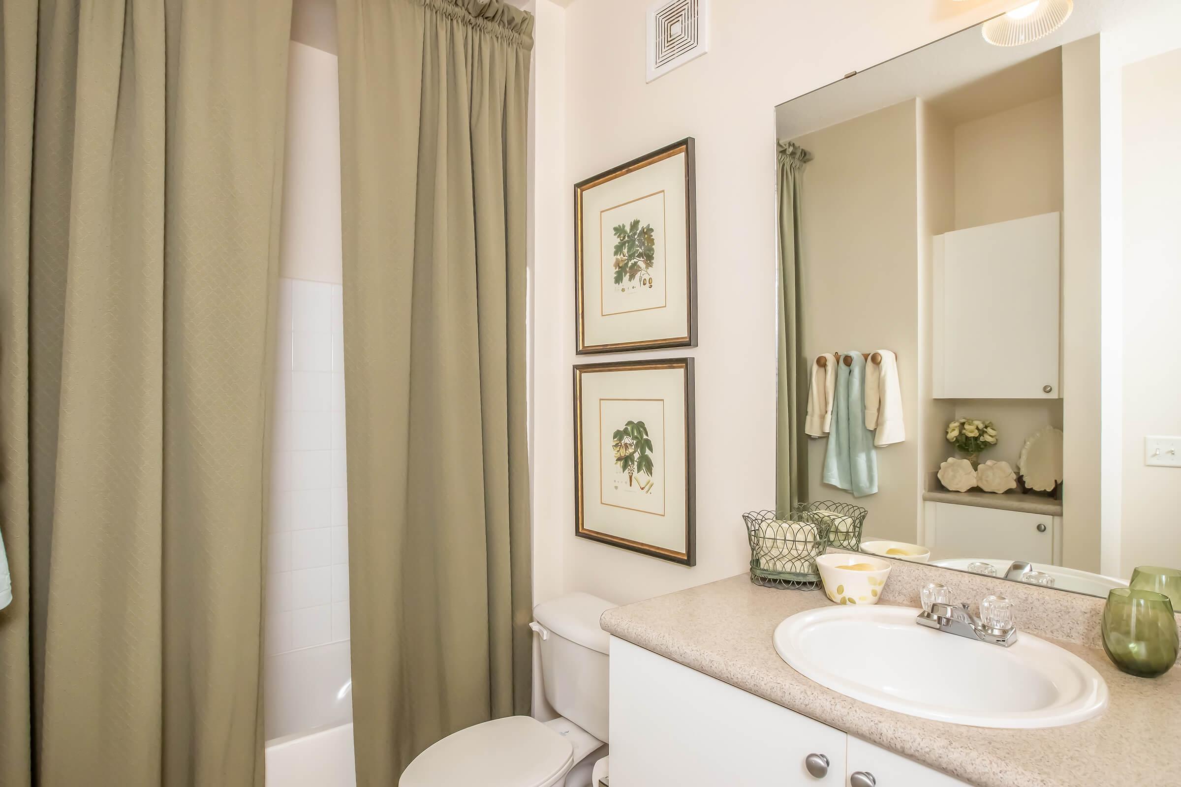 A well-lit bathroom featuring a white toilet, a sink with a countertop, and a mirror. The walls are painted a soft color, adorned with framed plant artwork. A light-colored shower curtain hangs in front of the bathtub, and towels are neatly arranged. A decorative basket and a floral arrangement add a touch of decor.