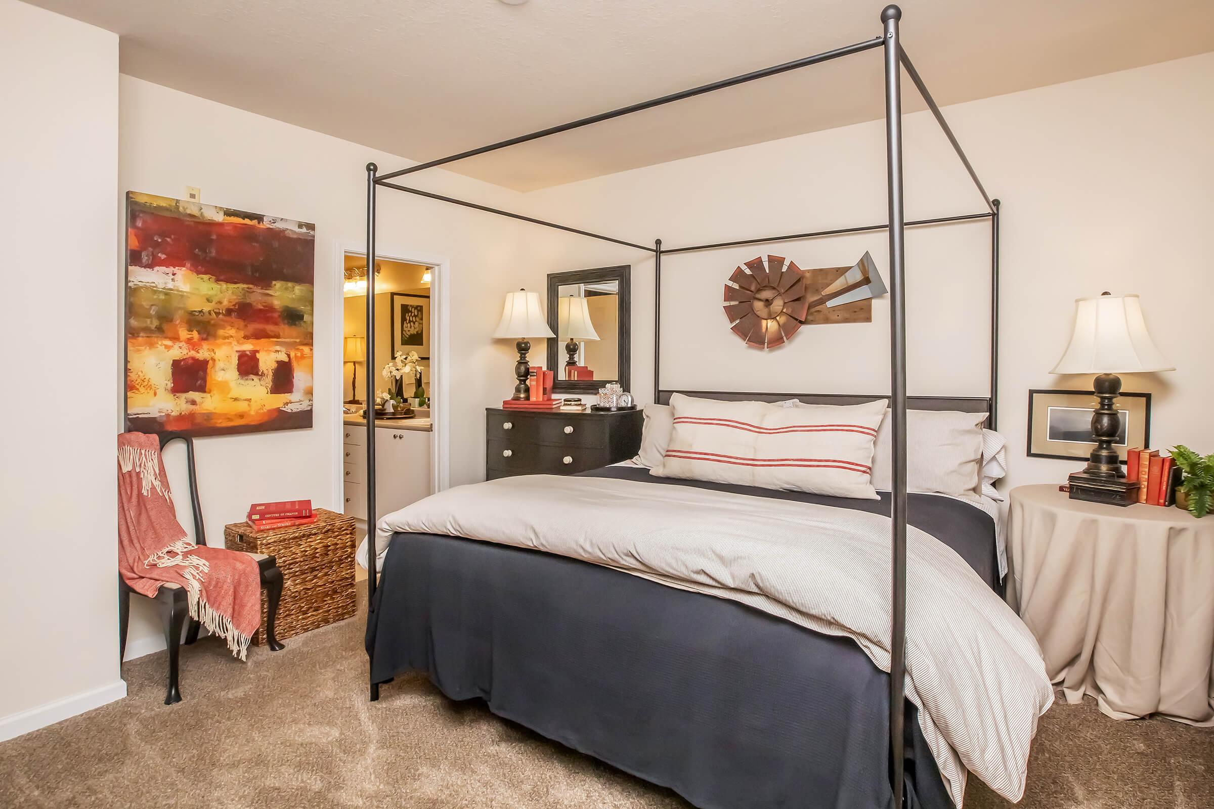 A cozy bedroom featuring a four-poster bed with gray bedding, a textured area rug, and bedside tables with lamps. A colorful abstract painting hangs on the wall, and a small accent chair sits nearby. The room is warm and inviting, with soft lighting and decorative touches that create a serene atmosphere.