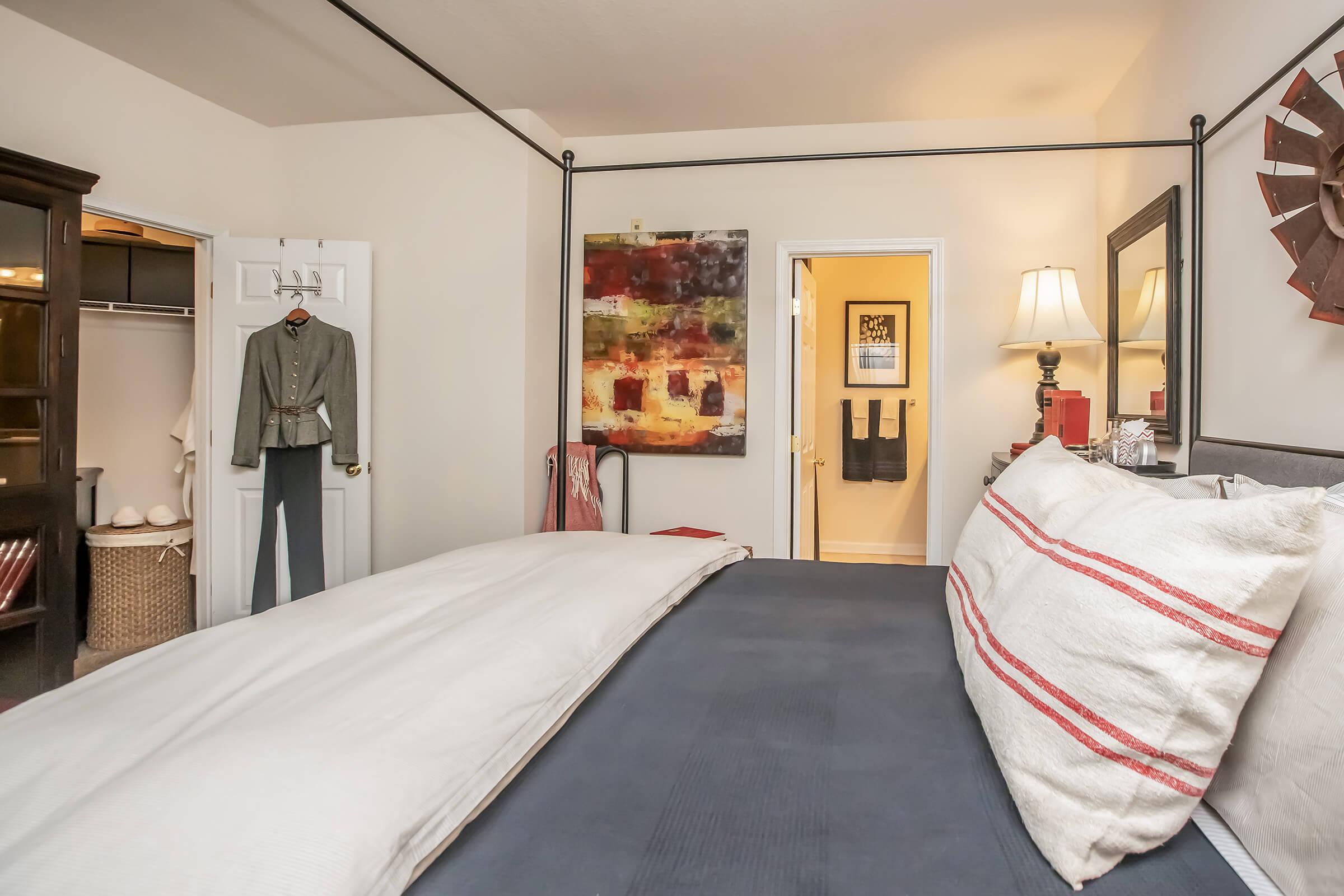 A cozy bedroom featuring a four-poster bed with white linens and red-striped accents. There's a modern artwork on the wall, a wooden wardrobe with clothing inside, and a hallway leading to another room. A lamp sits on a nightstand beside the bed, adding warmth to the space.