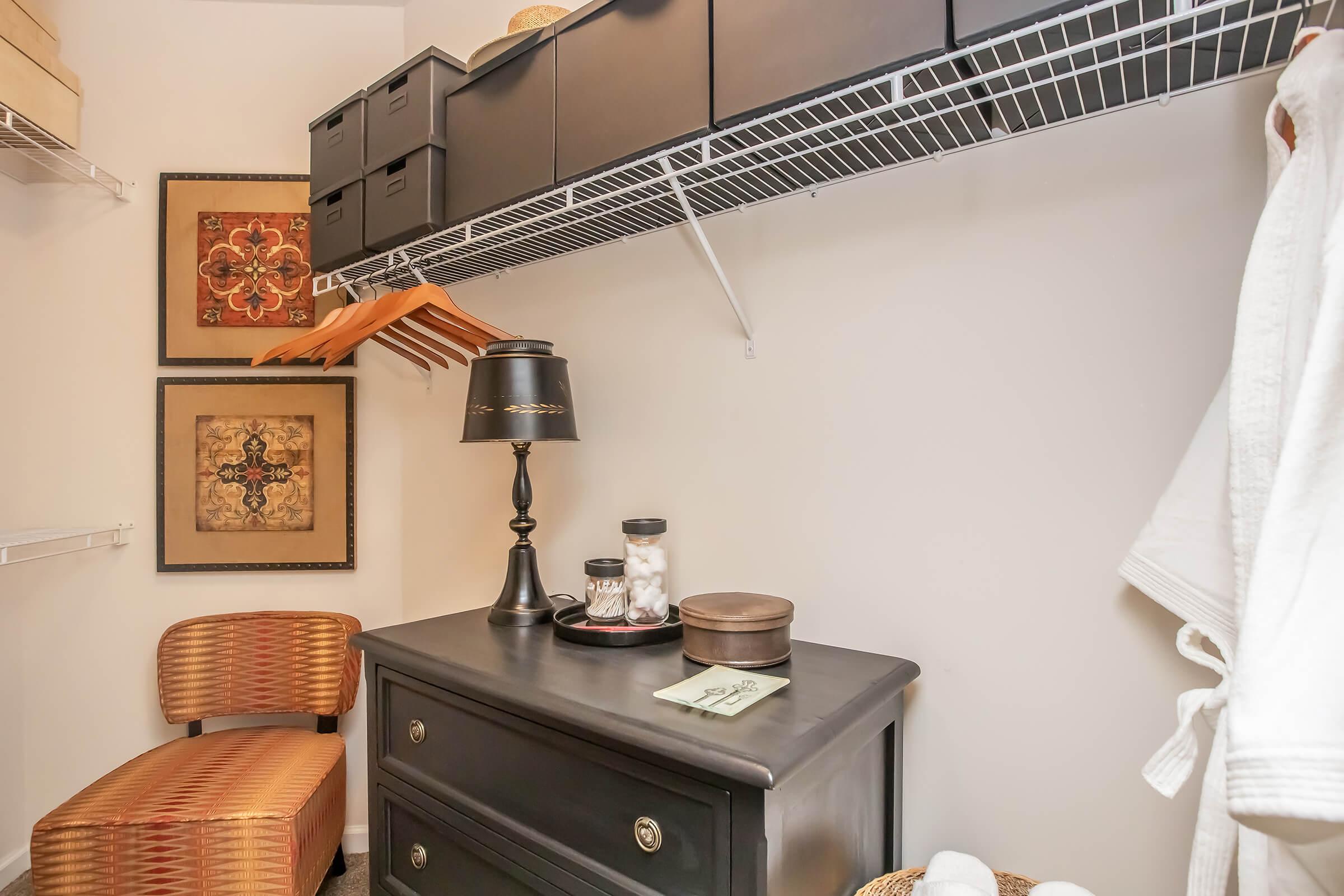A neatly organized closet featuring a black dresser, a decorative lamp, and several storage boxes on a wire shelf. There's a cozy chair, framed artwork on the wall, and wooden hangers displayed. The space appears tidy and inviting, ideal for storage and organization.