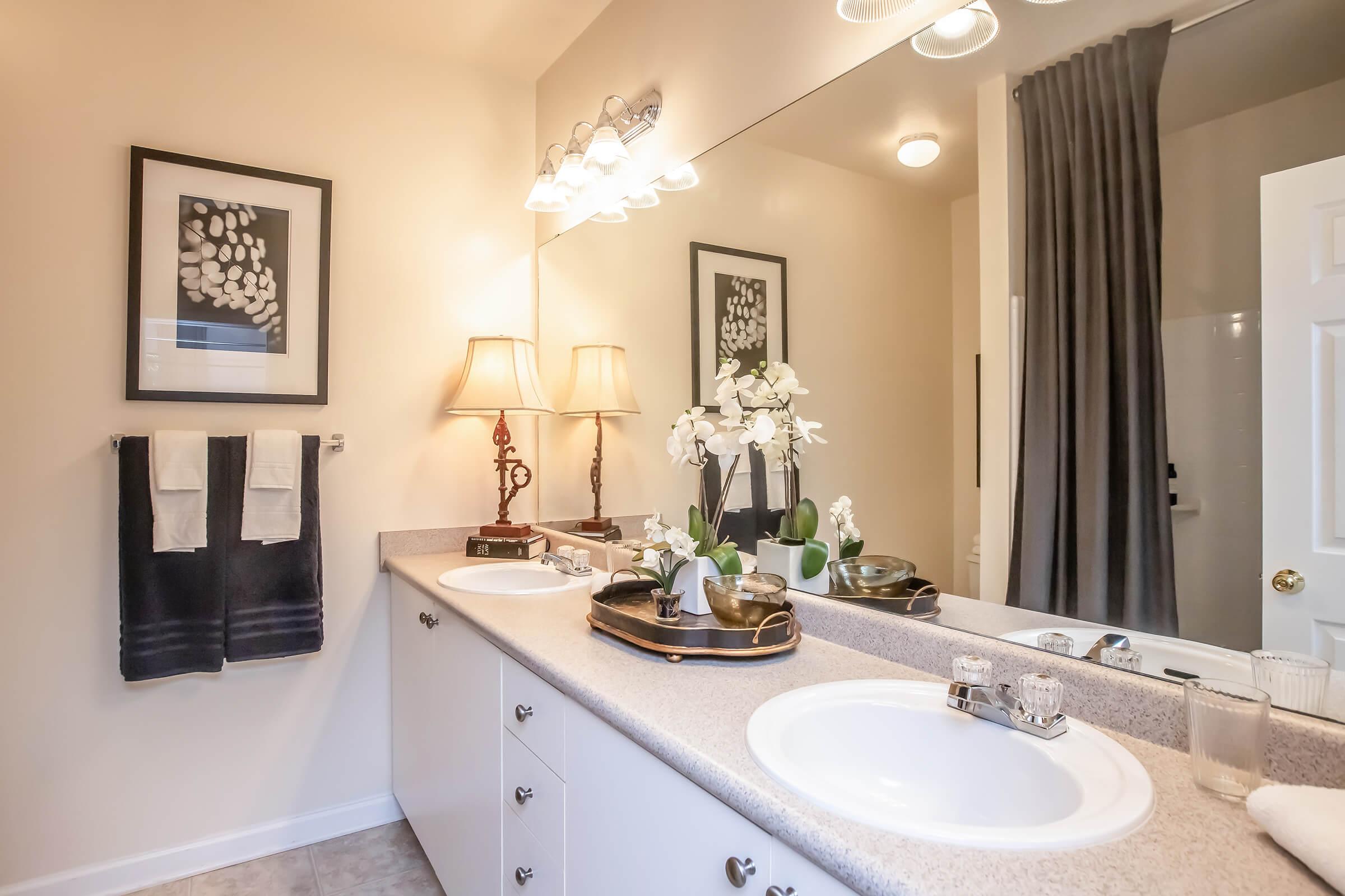 A modern bathroom featuring a double sink vanity with a light-colored countertop, two framed botanical prints on the wall, a lamp, and decorative items including a tray with toiletries. The mirror reflects the room's neutral tones, while dark towels hang on a rack nearby.