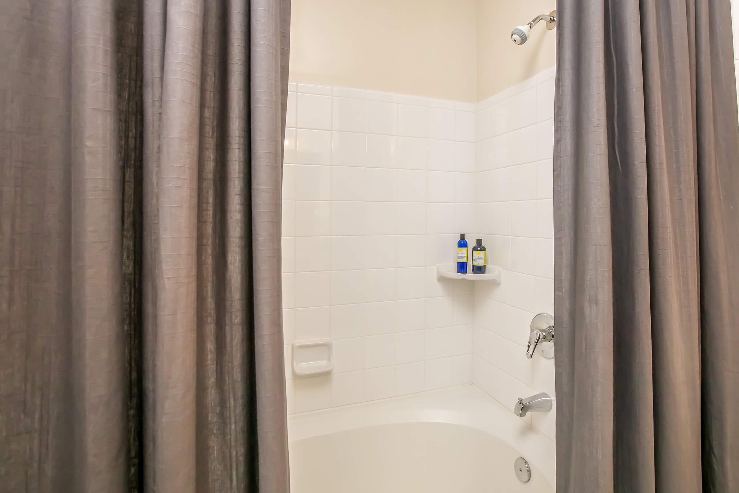 A bathroom with a shower area featuring a white tiled wall and a bathtub. Gray curtains are partially drawn to separate the shower from the rest of the bathroom. A small shelf holds two bottles of toiletries. The lighting is bright and neutral-colored, creating a clean and inviting atmosphere.