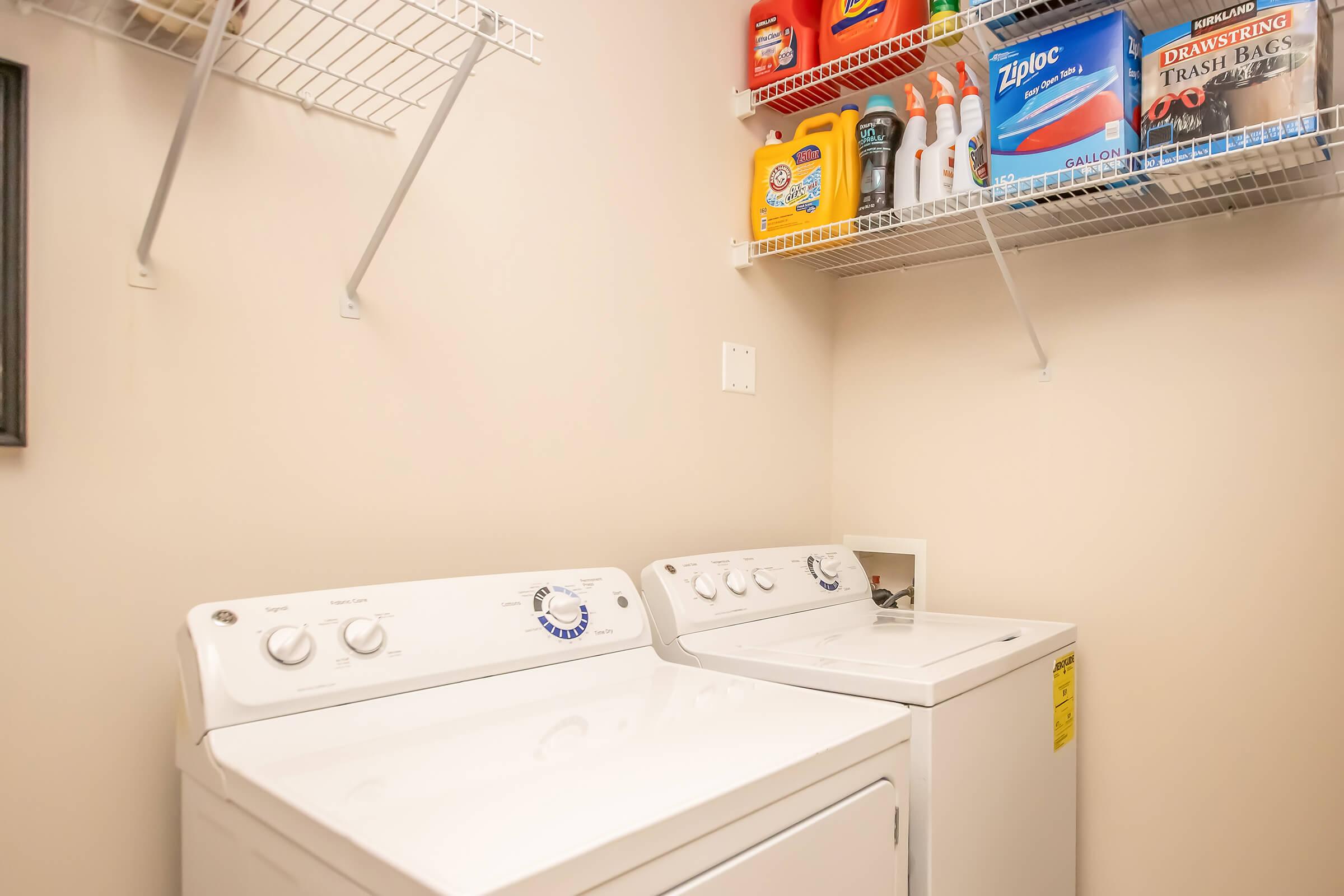 A small laundry room featuring a washer and dryer side by side. Above them, a wire shelf holds various cleaning supplies and laundry products, including detergent and trash bags. The walls are painted a neutral color, creating a clean and organized space.