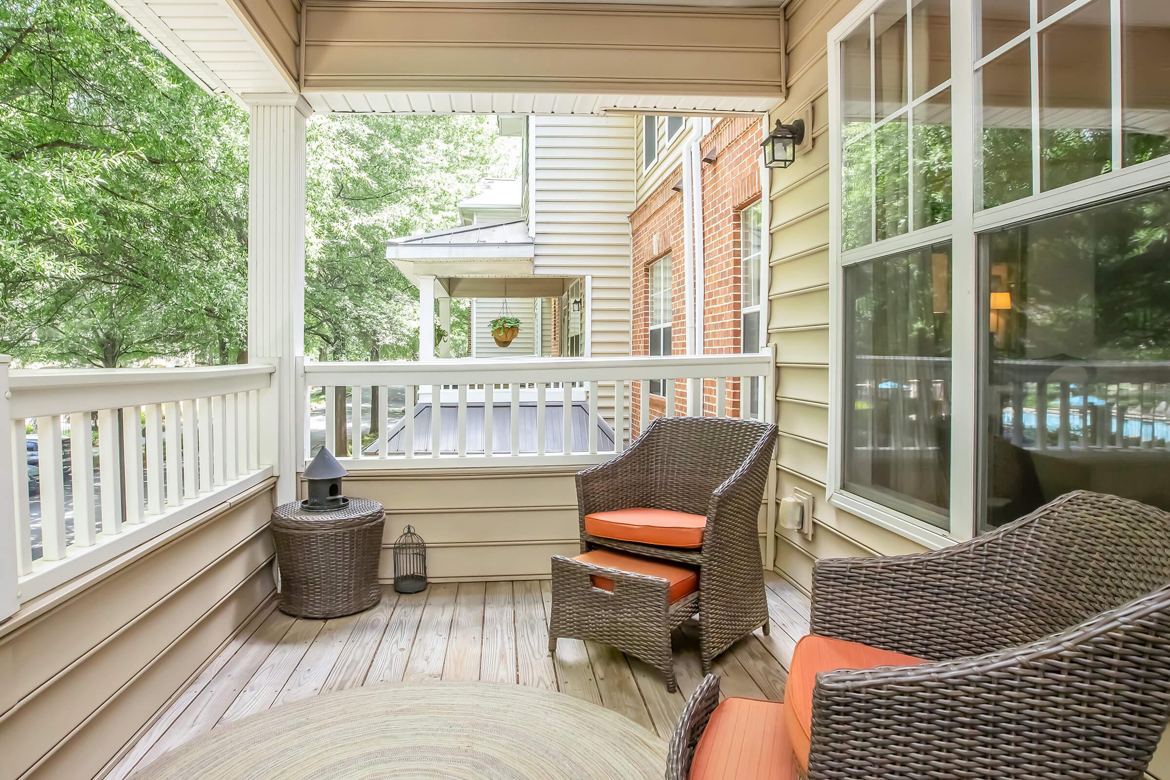 A cozy porch area featuring two wicker chairs with orange cushions, a round rug, and a small lantern on the floor. The space is surrounded by wooden paneling and features a railing with a view of greenery, creating a calm and inviting atmosphere.