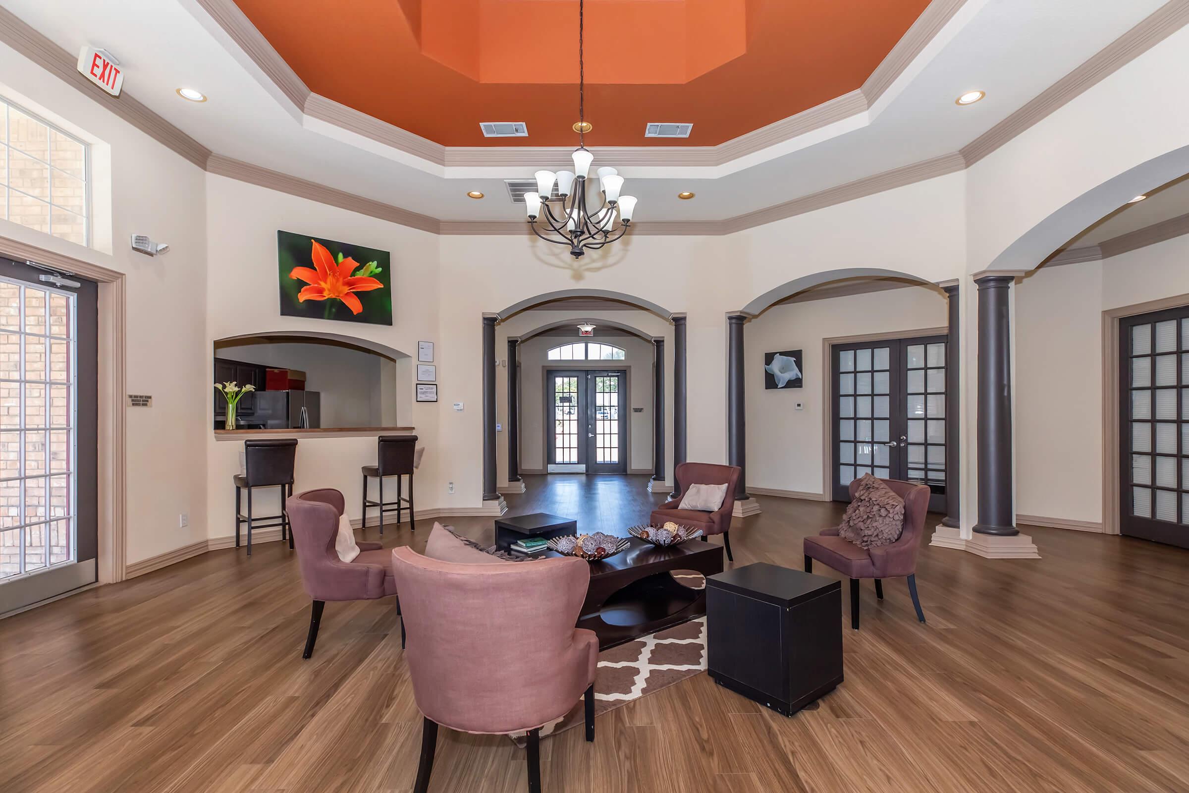 Interior view of a stylish lobby with high ceilings and an orange dome feature. Modern furniture includes plush chairs and a low coffee table. Decorative artwork is displayed on the walls, and there is a reception area in the background. The space is well-lit and features hardwood flooring.
