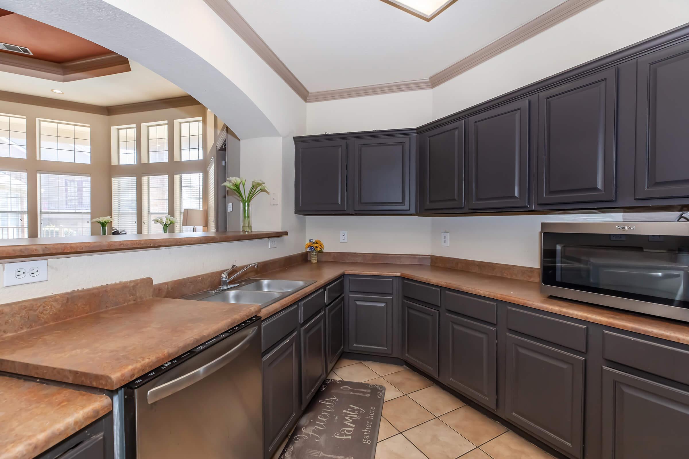 Modern kitchen with dark wooden cabinetry, a stainless steel sink, and a countertop. A built-in microwave is visible, along with decorative elements like a small flower arrangement. Natural light streams in through large windows, creating a bright and inviting atmosphere. Tile flooring complements the overall design.
