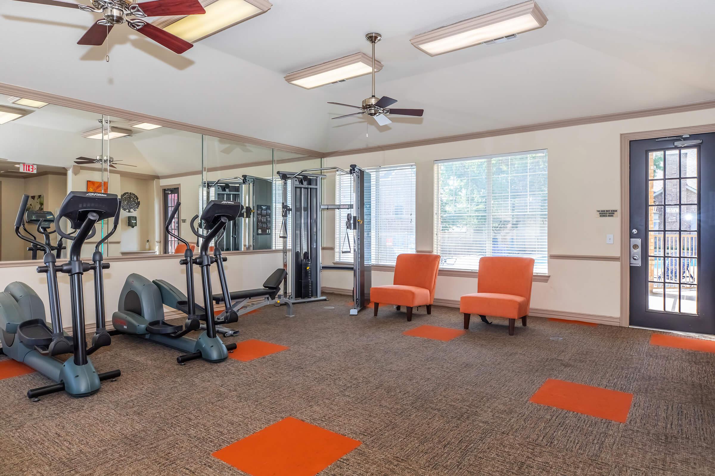 A well-lit gym area featuring exercise equipment like treadmills and elliptical machines, with two orange chairs by a large window. The space has ceiling fans and a carpeted floor with orange square accents. The atmosphere is inviting and designed for fitness and relaxation.