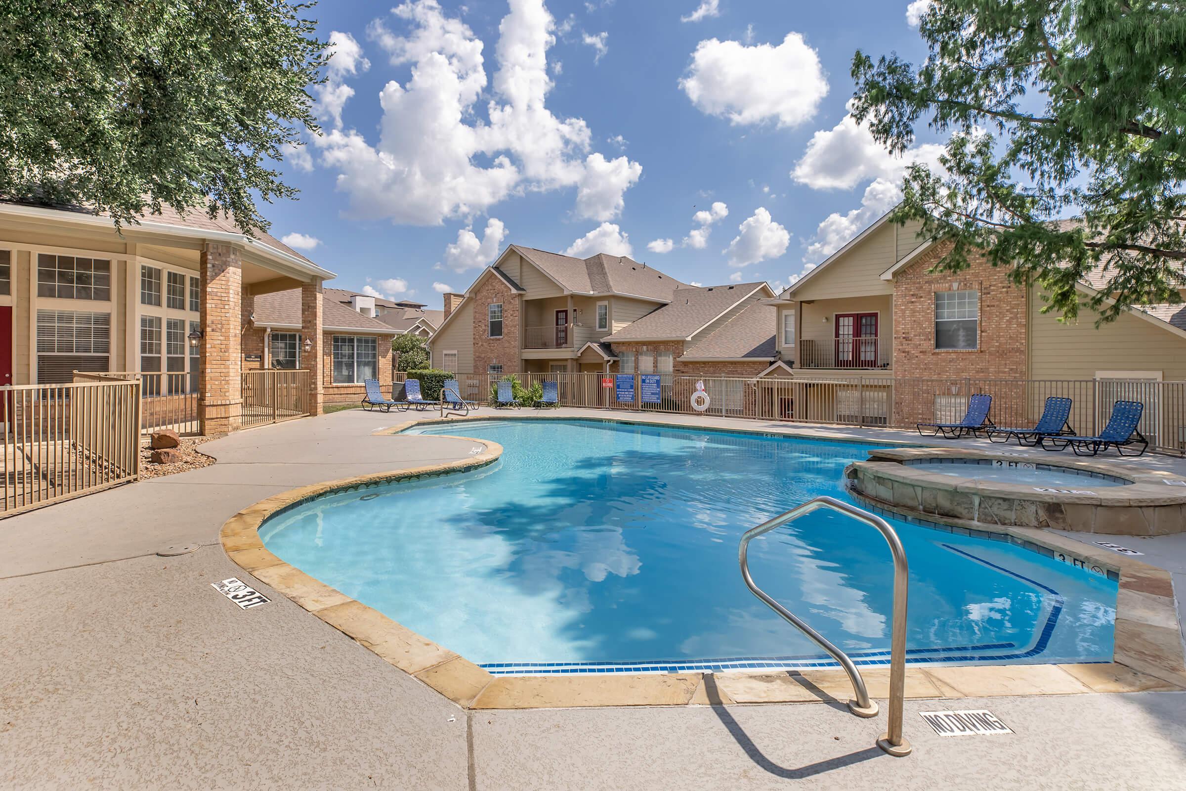 A clear, sunny pool area surrounded by apartment buildings. The pool has a gradual entry, with lounge chairs placed around it. Lush greenery and puffy white clouds in a bright blue sky complete the scene, creating a relaxing atmosphere.