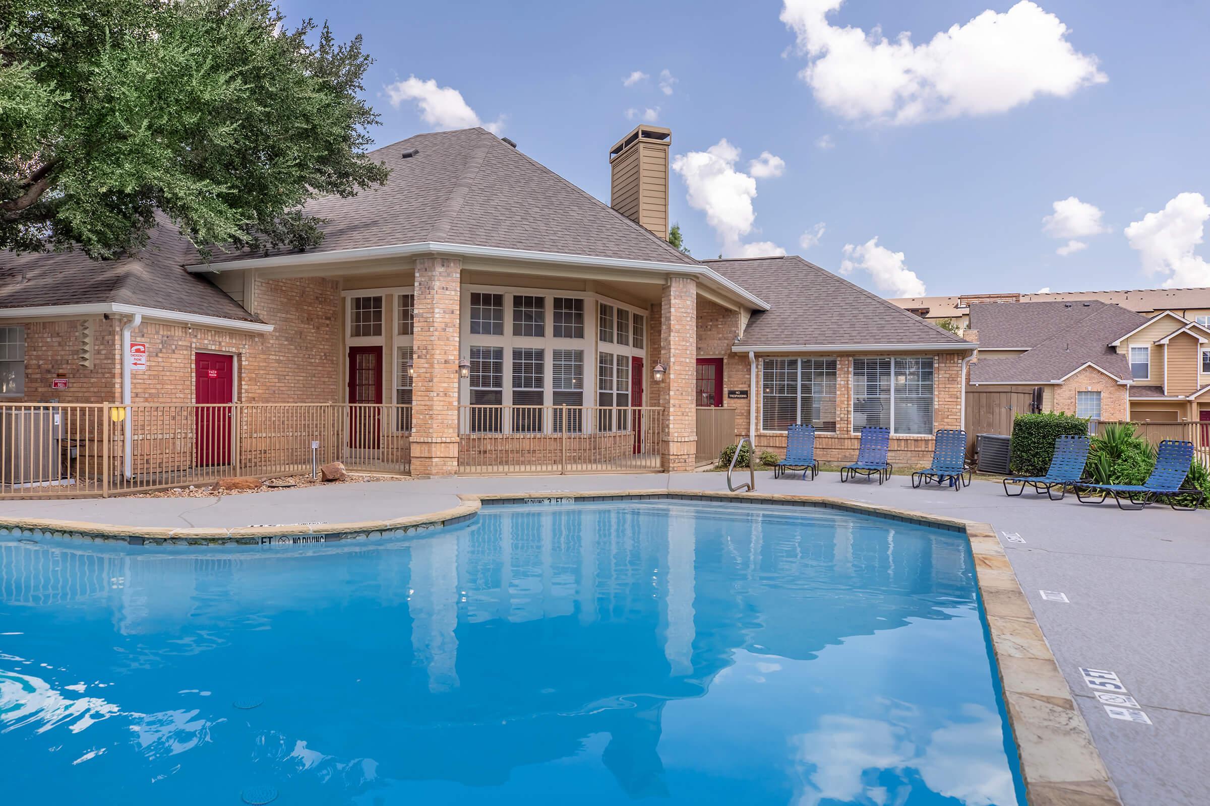 A well-maintained outdoor pool area featuring a clean blue pool surrounded by lounge chairs. In the background, there's a brick building with large windows and a sloped roof. Lush trees provide shade, and the sky is partly cloudy, creating a relaxing atmosphere.