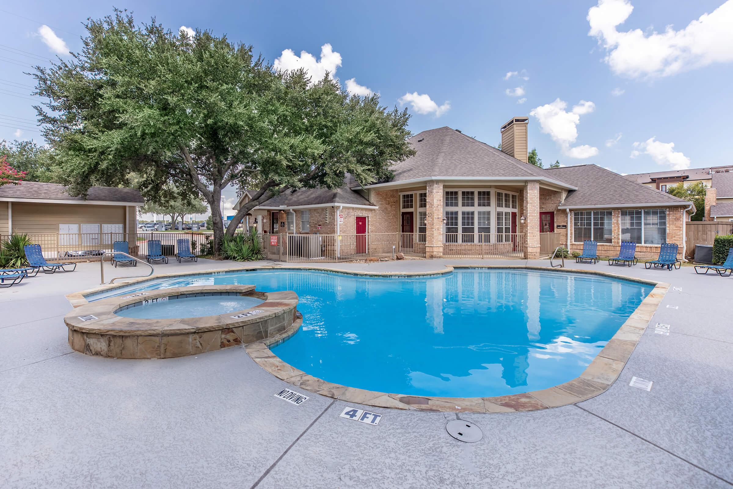 A clear blue swimming pool with a hot tub, surrounded by lounge chairs and trees. A light brown brick building with large windows and red doors is visible in the background. The scene is set under a sunny sky with some scattered clouds.