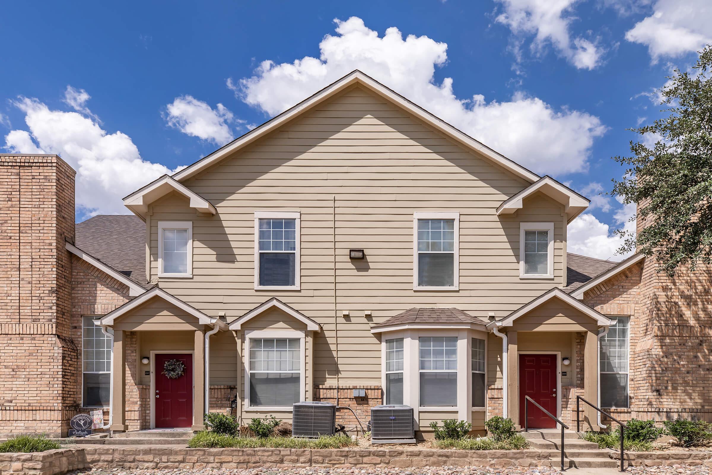 A two-story residential building with a beige exterior, featuring two front doors framed by windows. The architecture includes a gabled roof and stone accents. The front area is landscaped with a stone pathway, and the sky is bright blue with fluffy white clouds.
