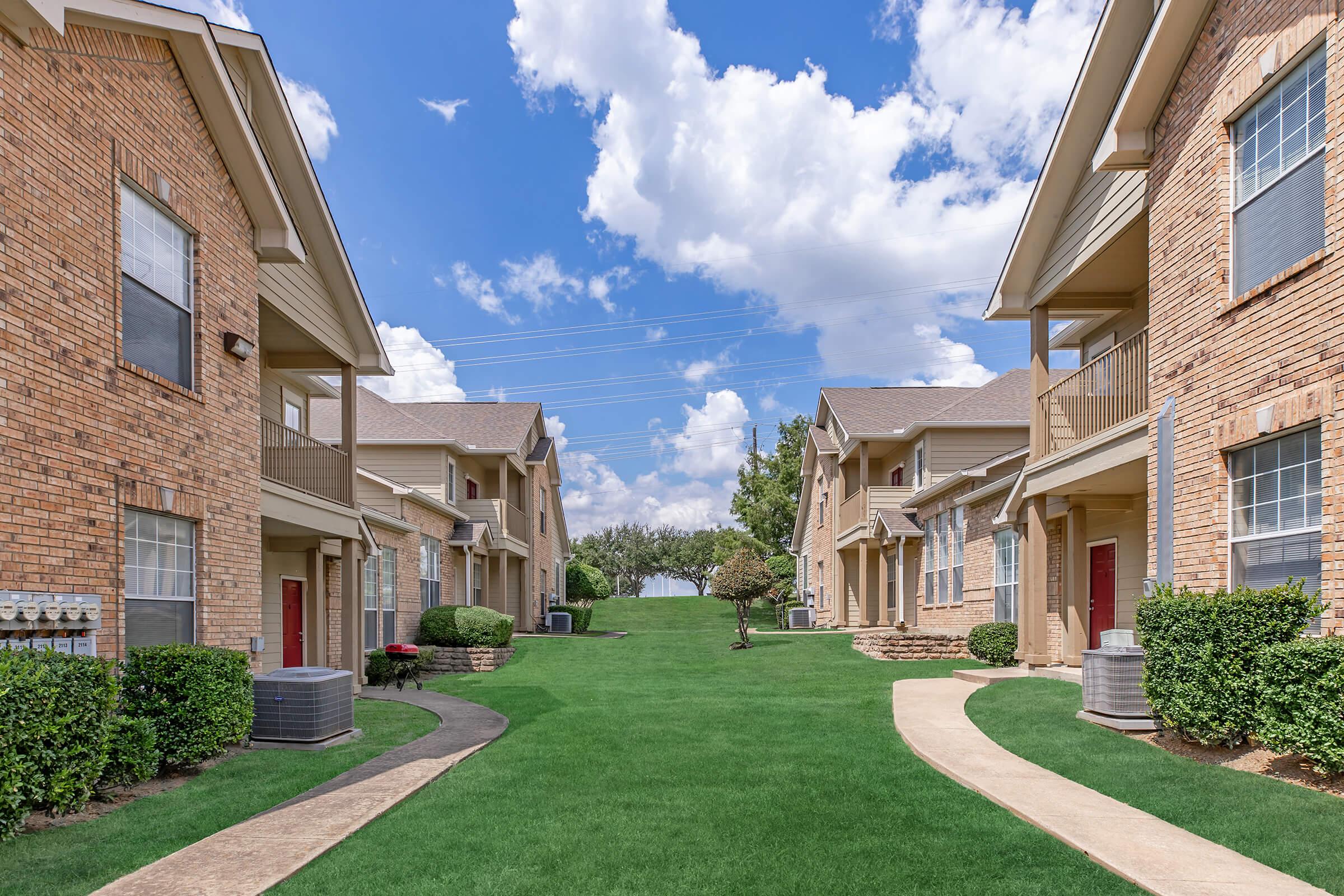 A view of a residential complex featuring two rows of beige brick apartment buildings with red doors. In between, a well-maintained green lawn with a pathway leading through the center, lined with small bushes and trees under a bright blue sky filled with fluffy white clouds.