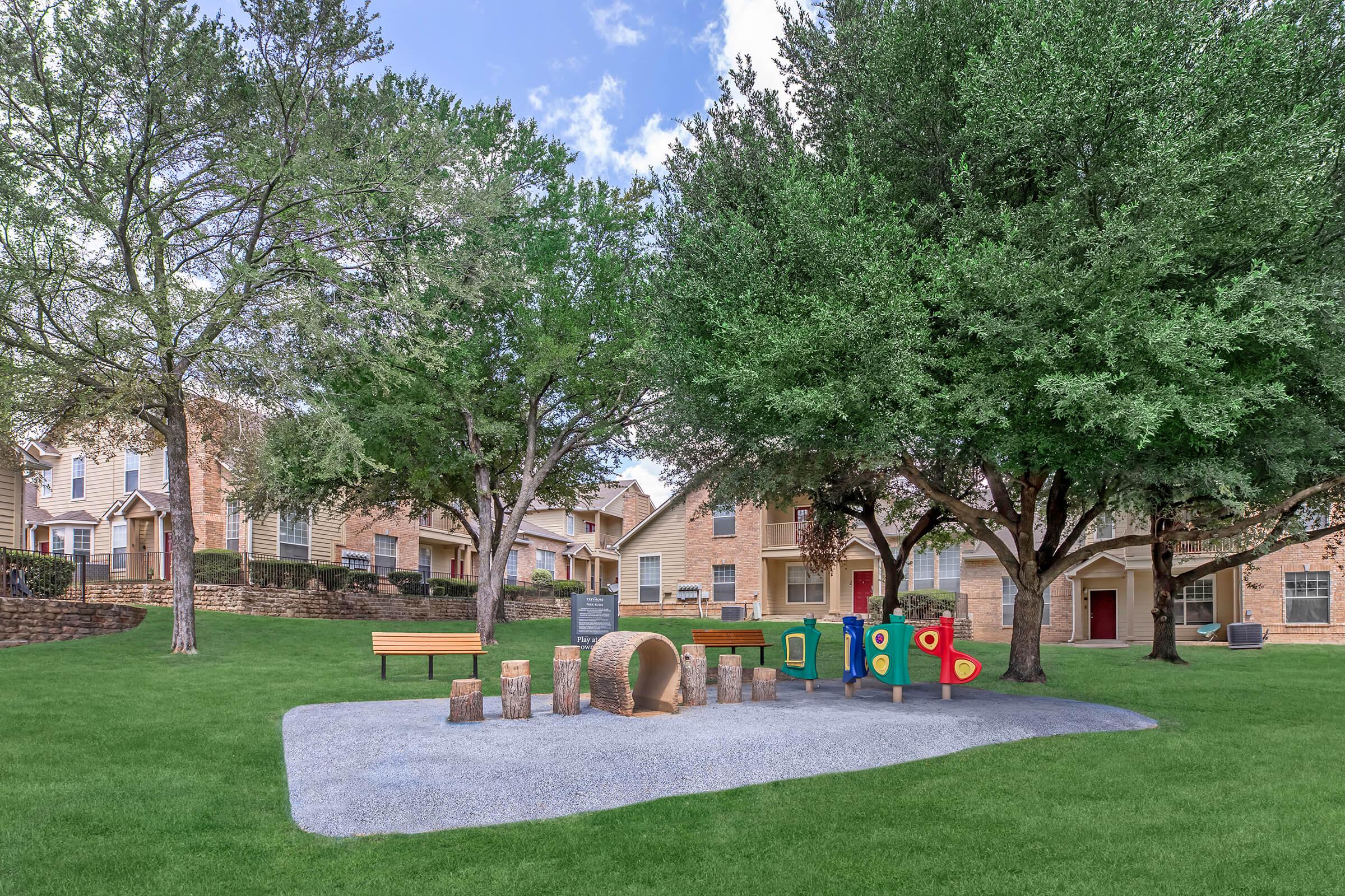 A playground area featuring colorful play structures and logs arranged for seating on a gravel surface. Surrounded by lush green grass and trees, the setting includes benches and residential buildings in the background, creating a bright and inviting outdoor space for children and families.
