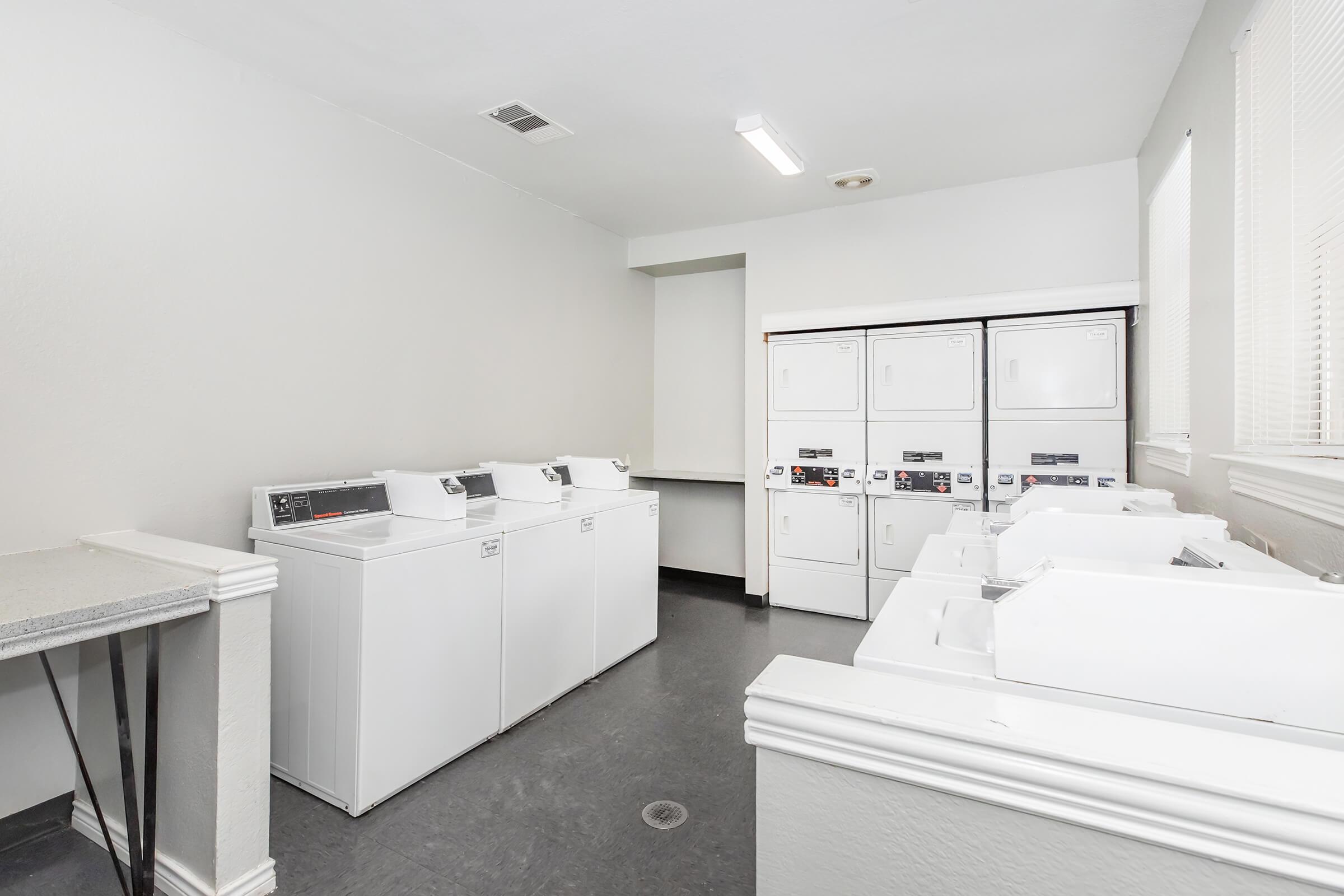 A clean and well-lit laundry room featuring several white washing machines and dryers against a gray wall. A countertop is visible on one side, and there are windows allowing natural light to enter the space. The room is tidy and organized, suitable for residential or communal laundry use.