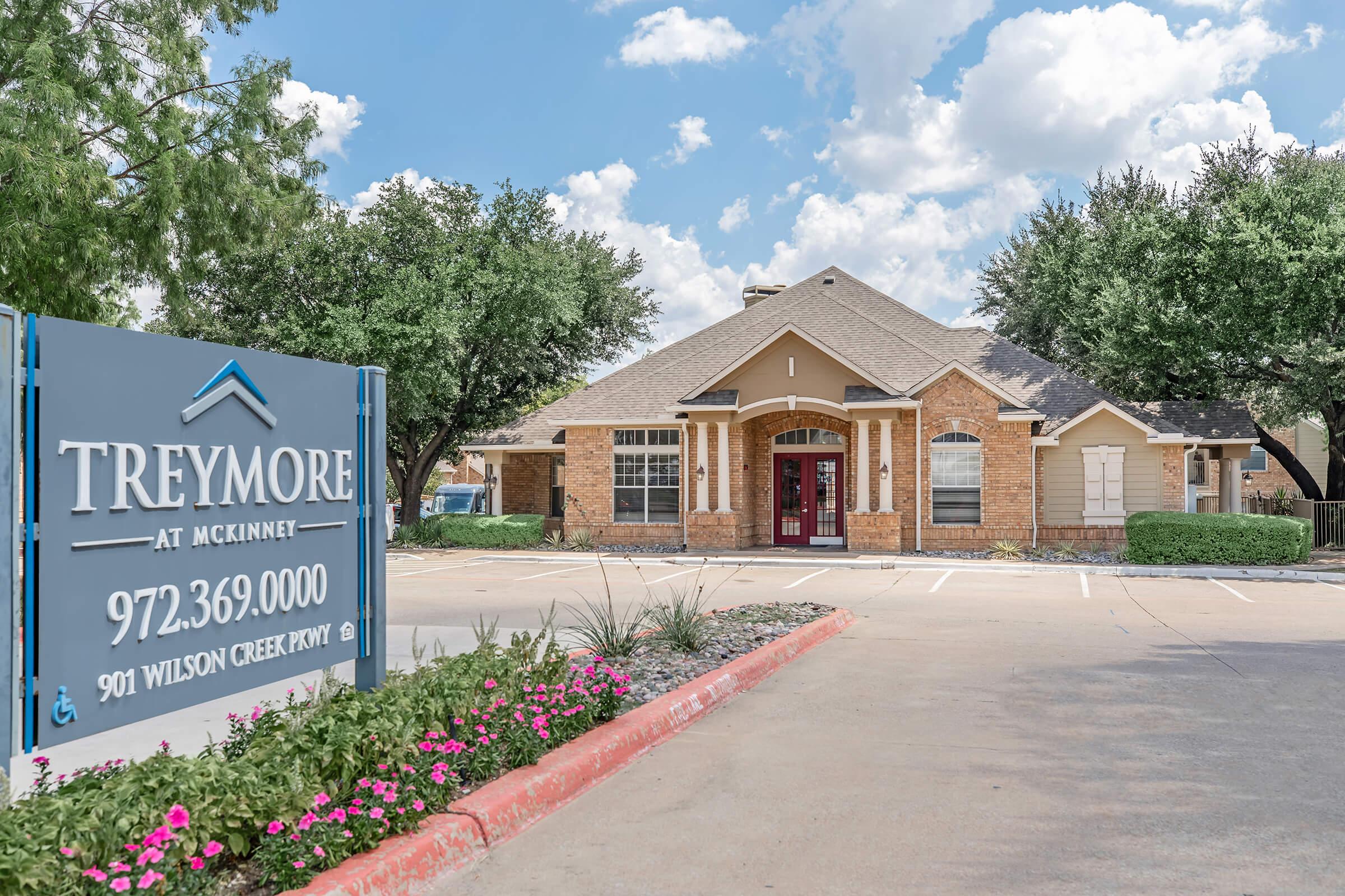 Exterior view of the Treymore at McKinney apartment complex, featuring a brick building with a peaked roof. The entrance is framed by landscaped areas with pink flowers and green shrubs. A large sign displays the name of the complex and contact information, under a blue sky with fluffy clouds.