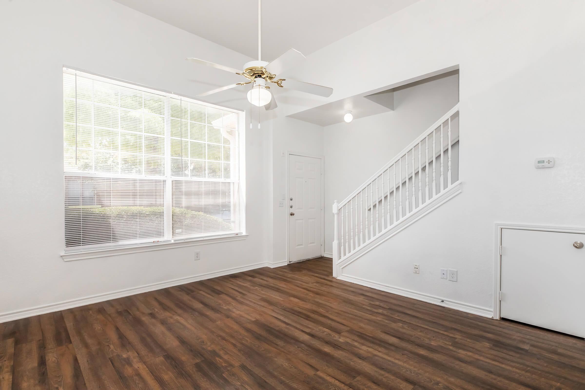 Interior of a bright and airy living room featuring a large window with blinds, light-colored walls, and a ceiling fan. A staircase is visible in the background, and the flooring is dark wood. The entrance door and a small white closet are also included in the scene.