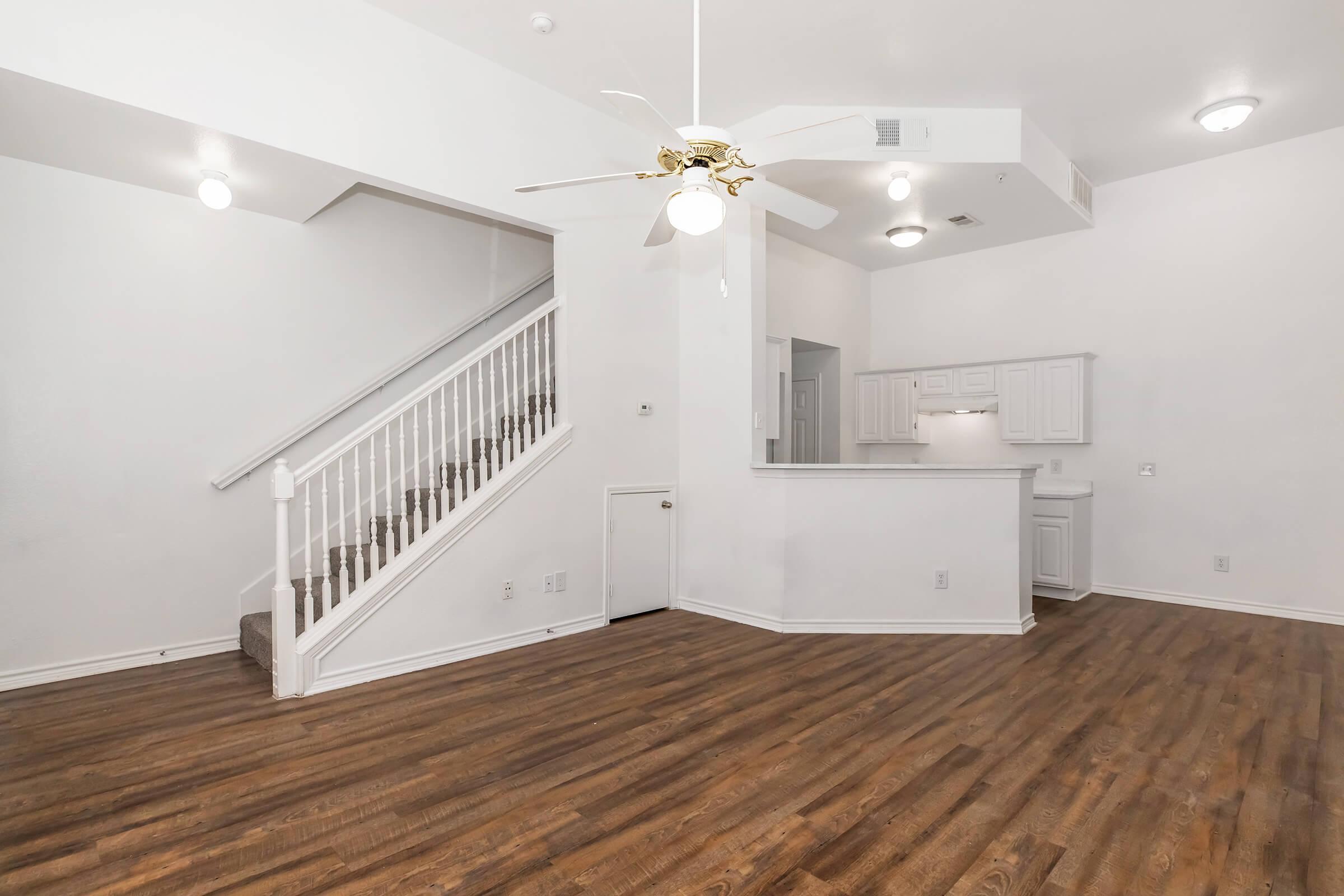 Spacious living area with hardwood floors, a ceiling fan, and a staircase leading to the upper level. The open layout includes a kitchen with white cabinetry visible in the background. Bright, natural light illuminates the room from multiple fixtures and windows.