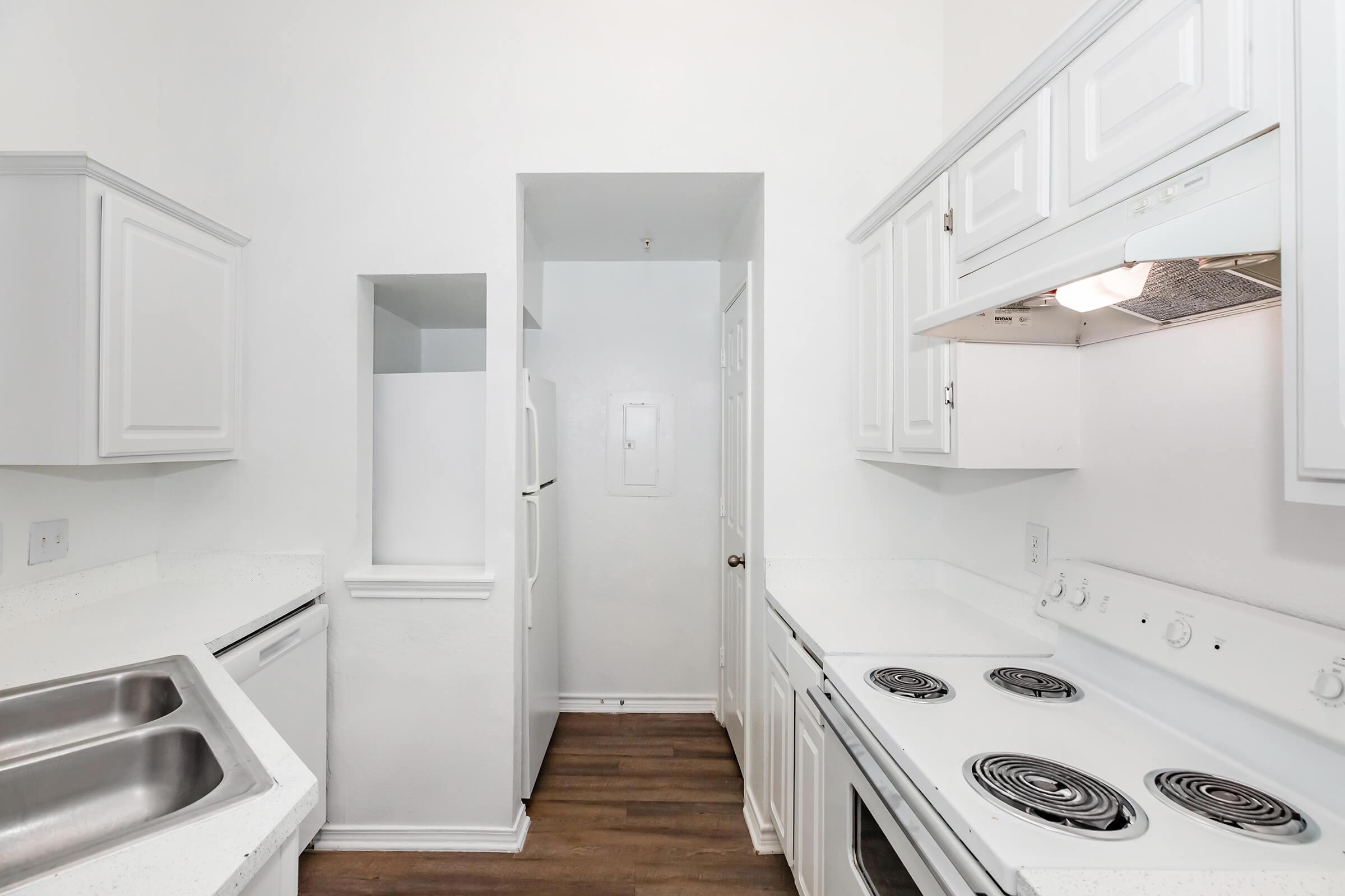A clean, modern kitchen featuring white cabinetry, a double sink, and a white stove with four burners. The space has a bright, minimalist design with light countertops, wooden flooring, and a doorway leading to another room.