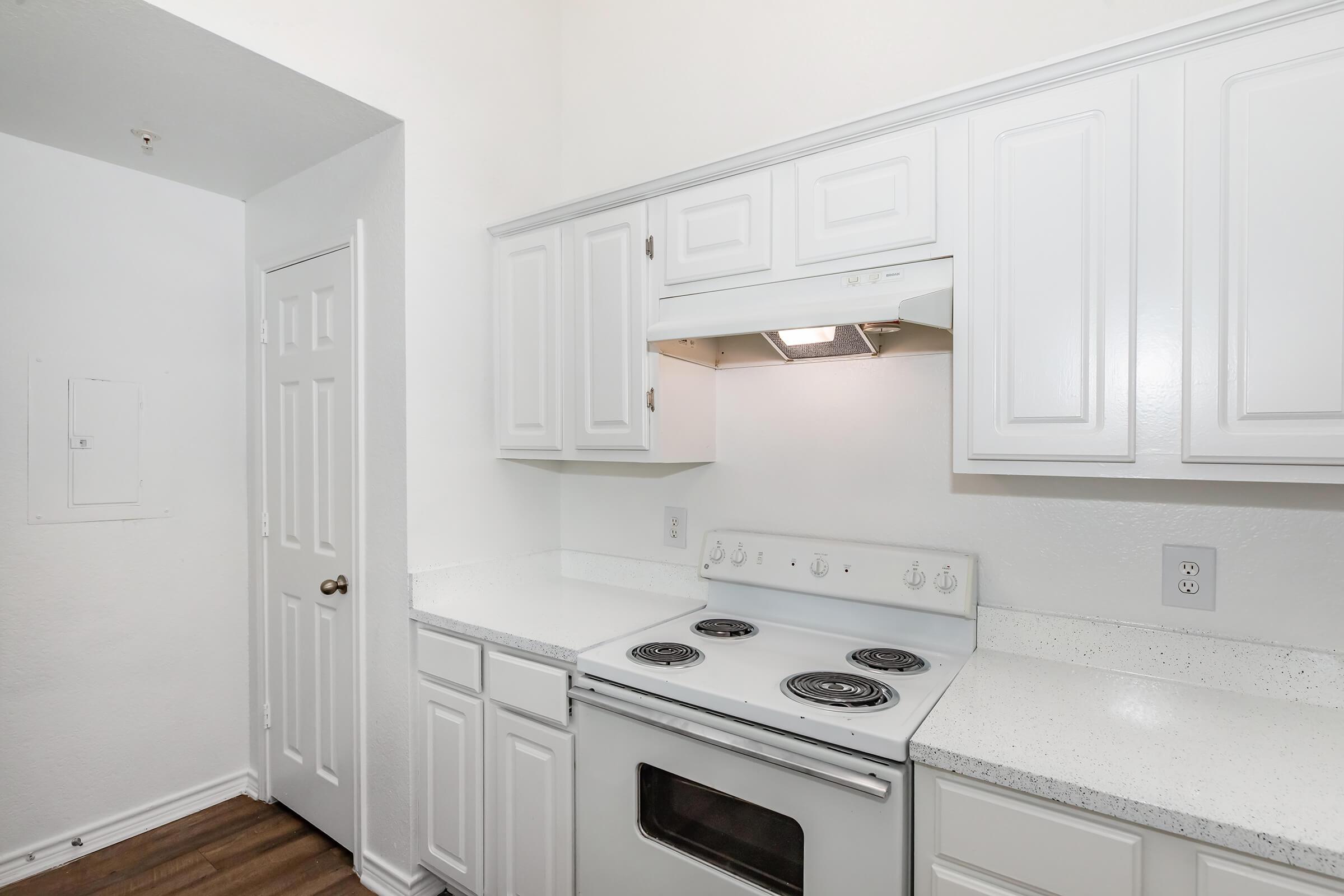 A small, modern kitchen featuring white cabinets, a white stove with four burners, and a countertop. The walls are painted white, and there is a door leading to another room. The overall aesthetic is clean and minimalist.