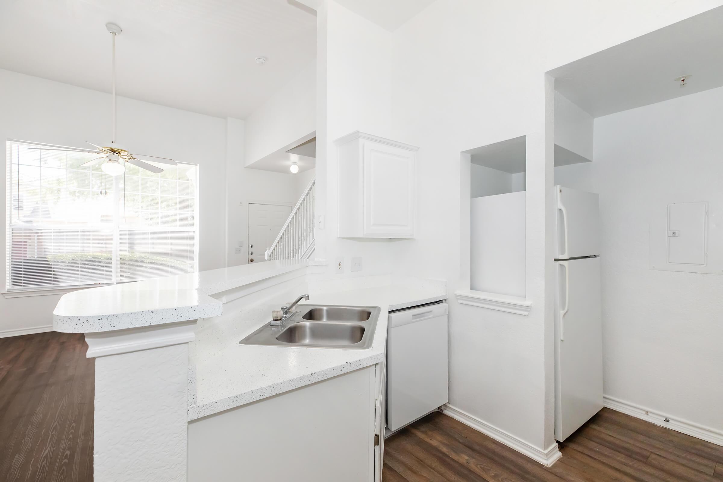 A modern, bright kitchen featuring white cabinetry, a double sink, and a dishwasher, with a refrigerator nearby. The open layout connects to a well-lit living area with large windows. The flooring is a warm wood laminate, creating a cozy atmosphere.
