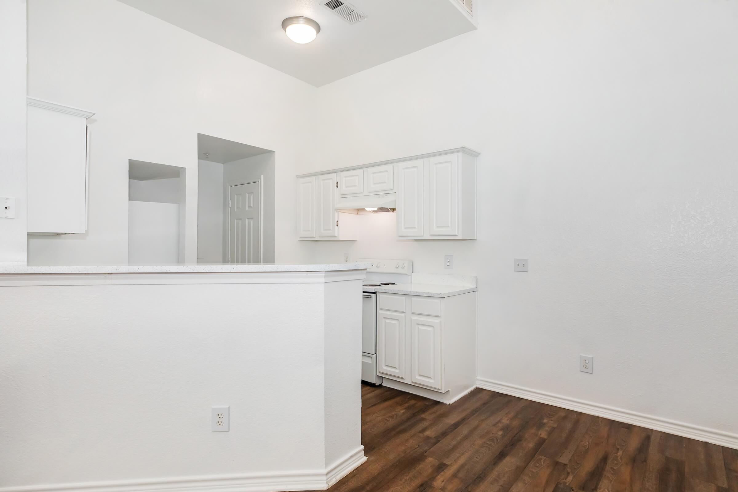 A bright and spacious kitchen area with white cabinets, a stove, and light-colored walls. The flooring is dark wood, adding contrast to the overall light aesthetic. The layout appears open, promoting a welcoming atmosphere.