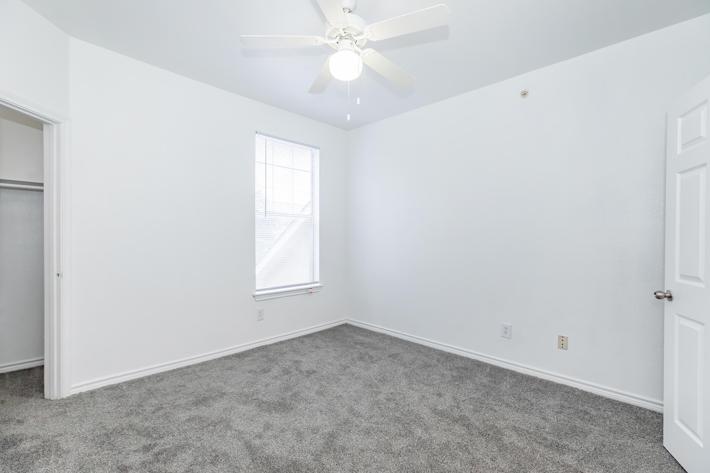 A bright, empty room with light gray carpet and white walls. The ceiling features a ceiling fan, and a closed door is visible on one side leading to a closet. A window allows natural light to enter, creating a spacious feel.