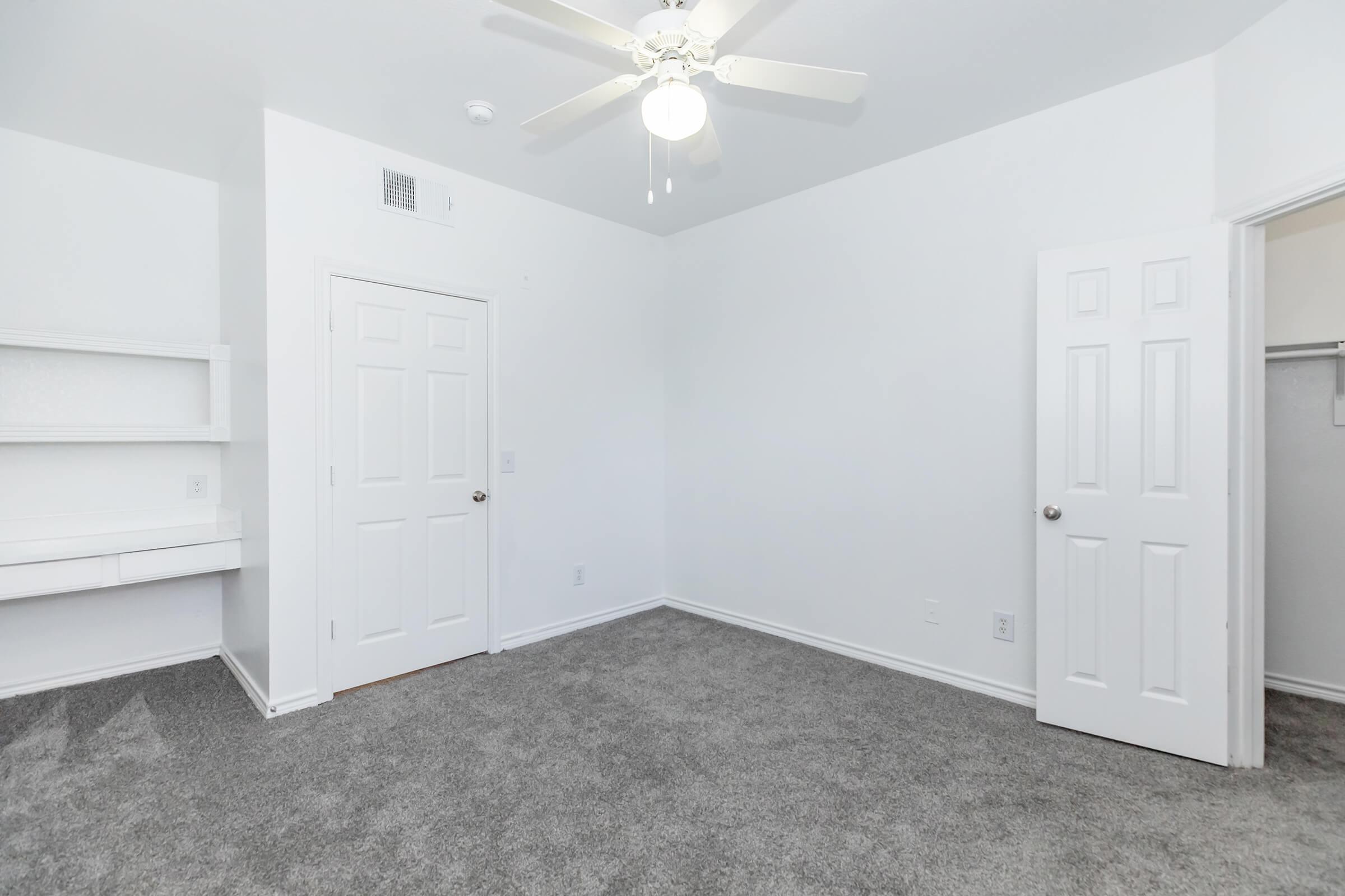 A vacant room with light gray carpet and white walls. It features a ceiling fan, two closed doors on the right leading to a closet and another area, and a small shelf mounted on the left wall. The room is brightly lit, emphasizing its spacious and clean appearance.