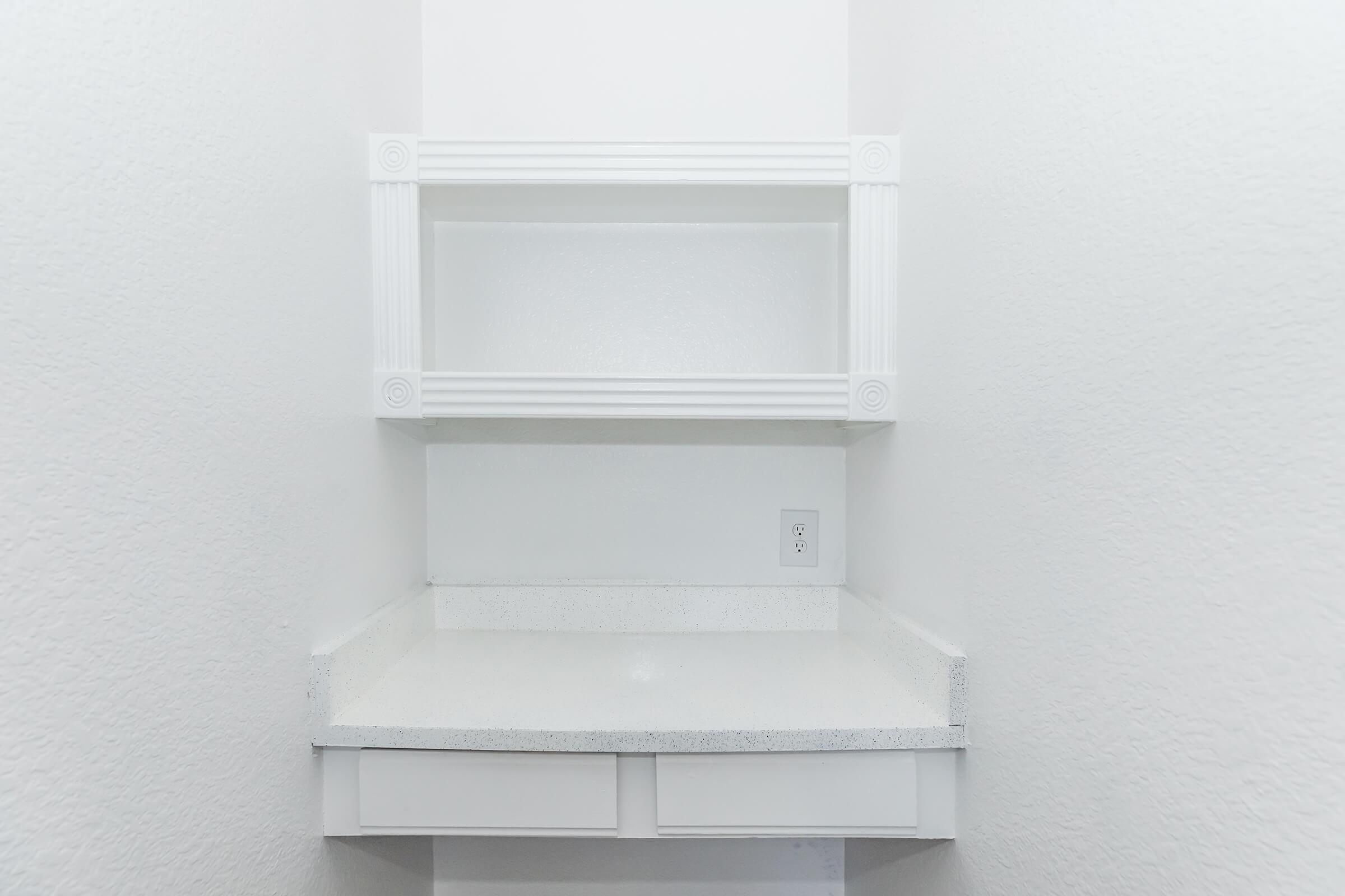 A clean, minimalist space featuring a white wall with a small shelf at the top. Below the shelf is a smooth, white countertop with a drawer beneath it. The overall design is simple and modern, with a bright and airy feel due to the light colors.