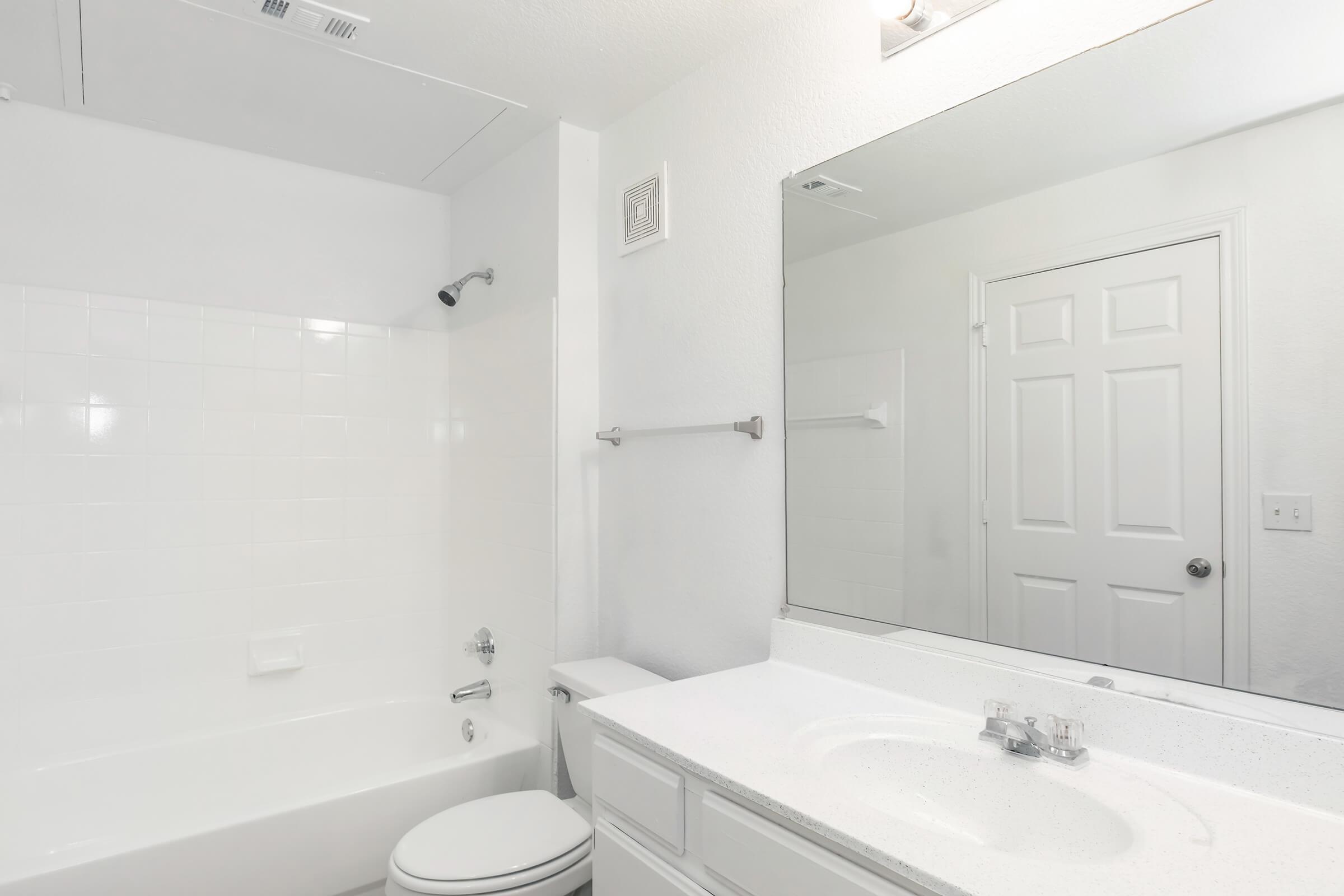 A clean, modern bathroom featuring a white bathtub, a wall-mounted showerhead, and a large mirror above a white sink with a countertop. The walls are painted white, enhancing the bright and spacious feel of the room. A closed door is visible in the background.