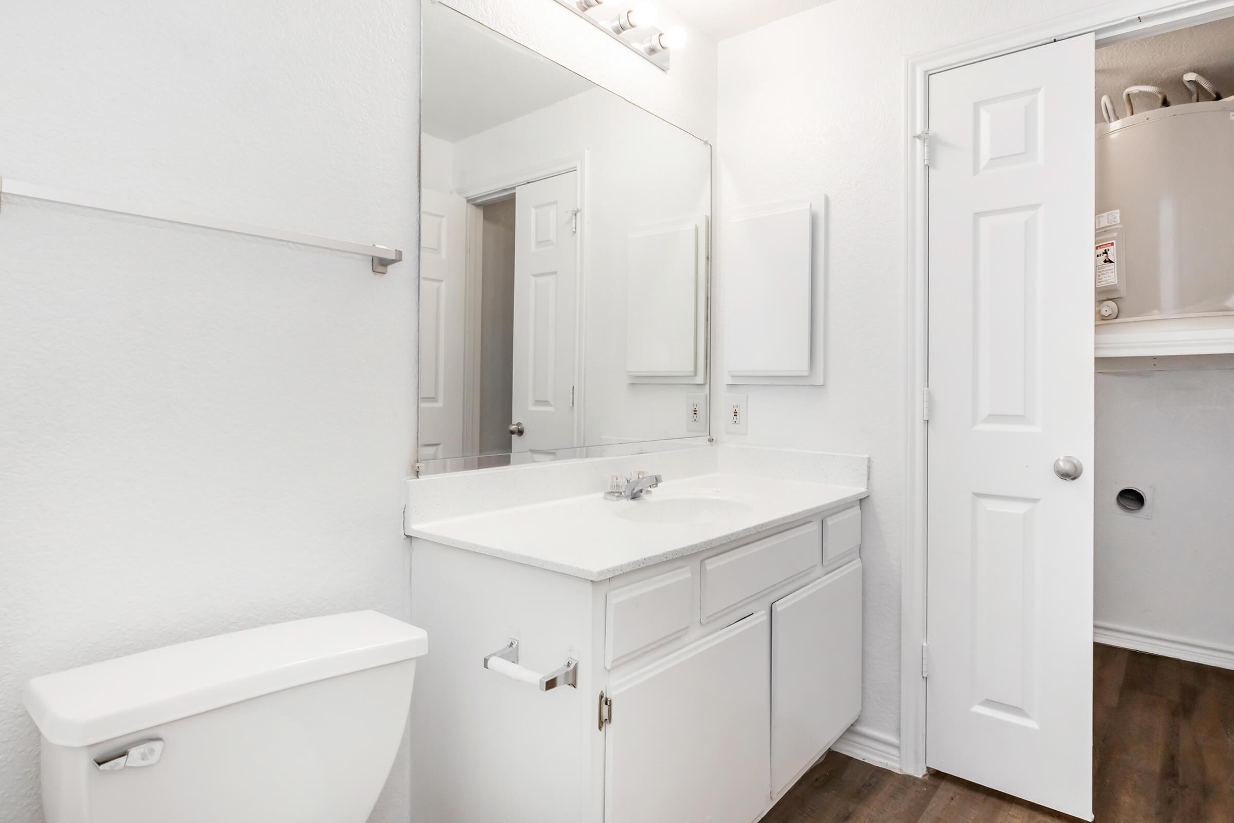 Bright and modern bathroom featuring a white vanity with a sink, a large mirror, and a toilet. The walls are painted in a light color, and there is a door leading to a closet or laundry area. The flooring is wood-like, adding a warm touch to the space. Overall, it has a clean and inviting appearance.