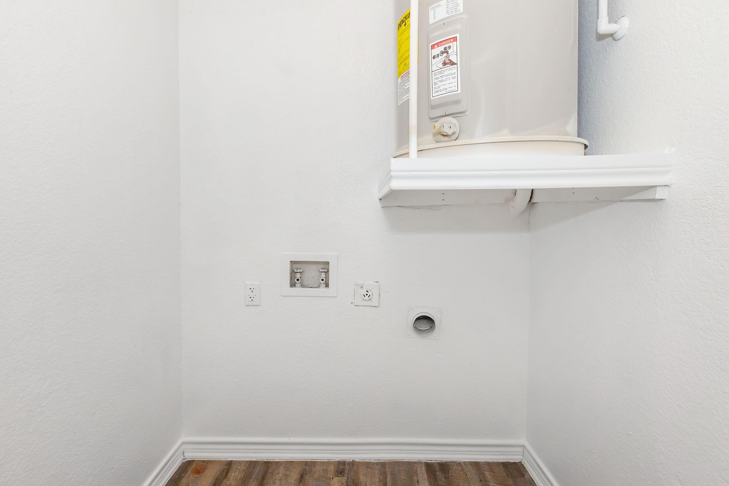 A small, empty laundry room with plain white walls, featuring a water heater mounted on the wall. Electrical outlets and a ventilation pipe are visible on the wall, along with a light-colored wooden floor. The space is uncluttered and tidy, suggesting it is designed for practical use.