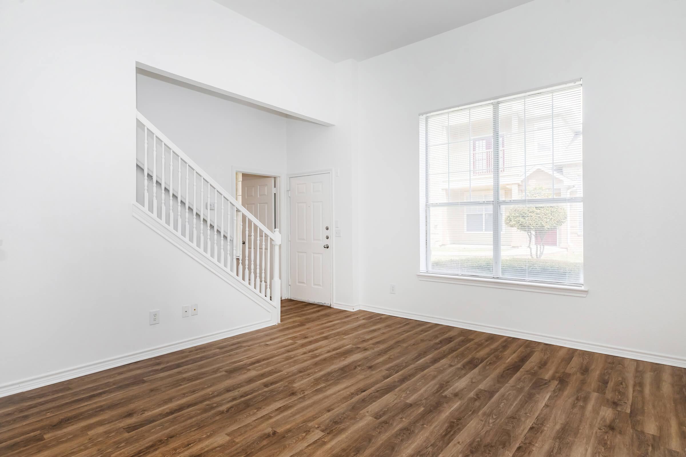 A spacious, empty room featuring light-colored walls and wooden flooring. A staircase is visible to the left, while a large window allows natural light to fill the space. The door and window overlook a garden or exterior area, creating a bright, airy atmosphere.