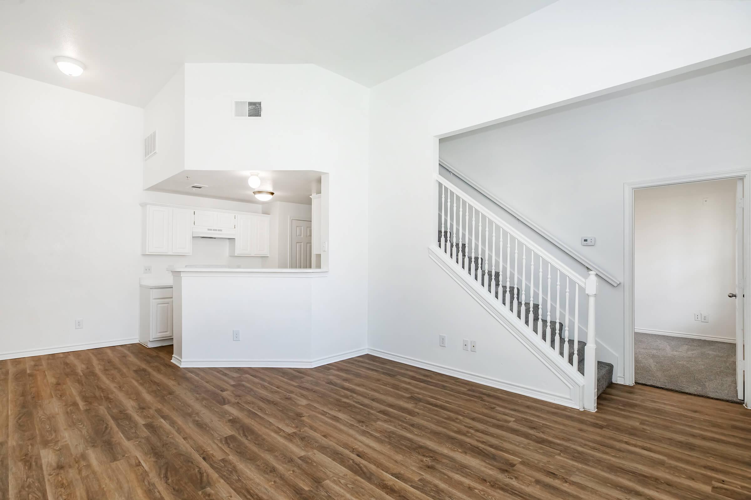 A spacious living area features light-colored walls and hardwood floors. A staircase leads to the upper level, while an open kitchen with white cabinetry is visible in the background. Natural light streams in from windows, creating a bright and airy atmosphere. An empty corner suggests potential for furniture placement.