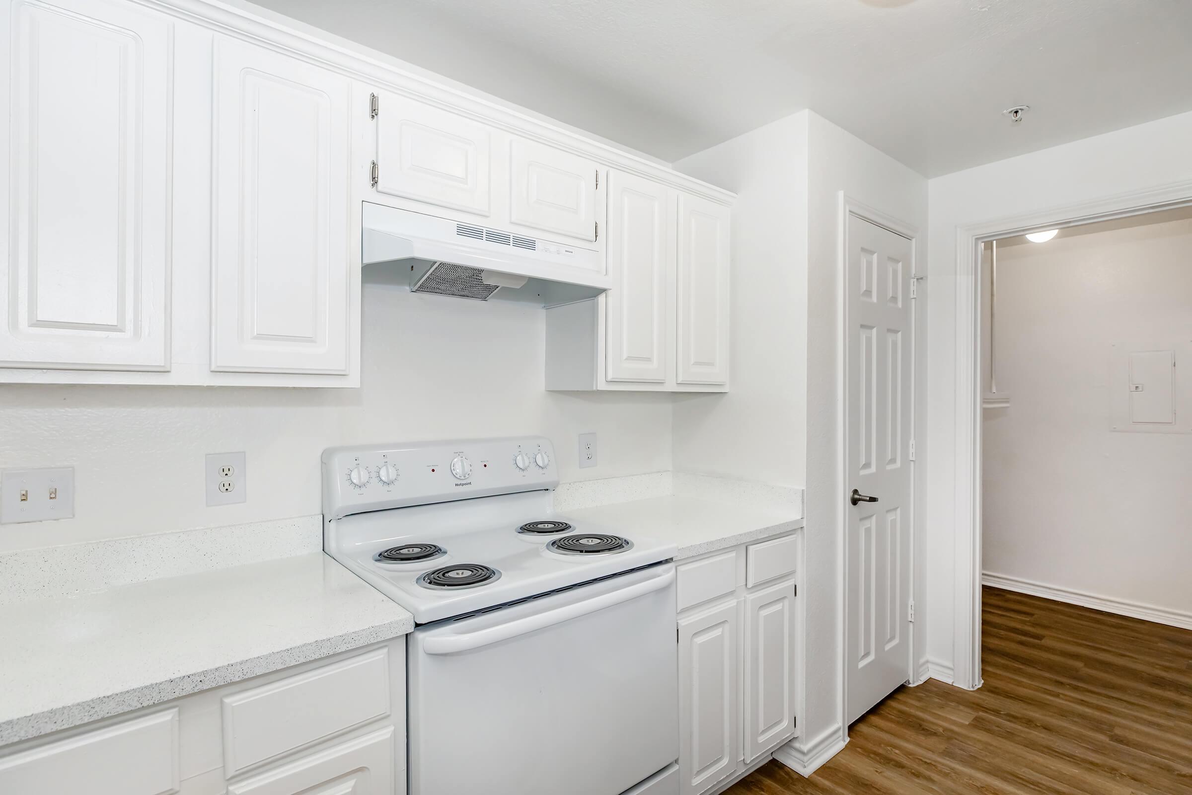 Bright, modern kitchen featuring white cabinetry, a stove with four burners, and a vent hood. The countertops are light-colored and clean, complemented by a wood floor. A doorway leads to another room, adding to the spacious feel.