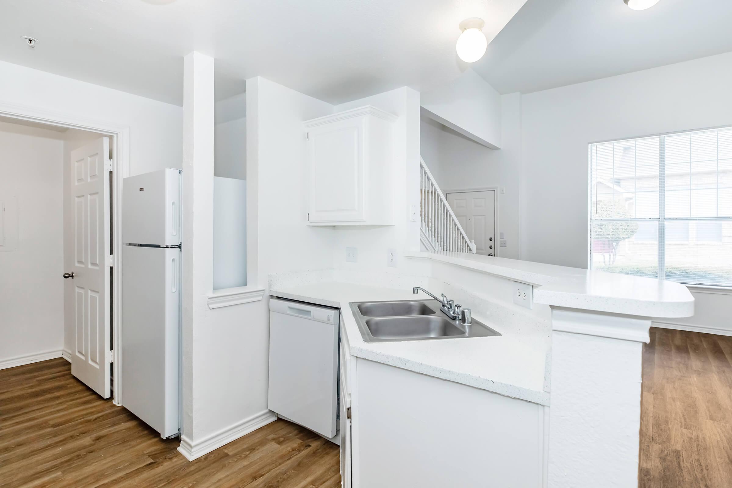 A modern kitchen featuring white cabinets, a sink, and stainless steel appliances. There’s a dishwasher next to the sink and a refrigerator nearby. The room has light-colored walls, ample natural light from a large window, and wooden flooring, creating a bright and spacious atmosphere.