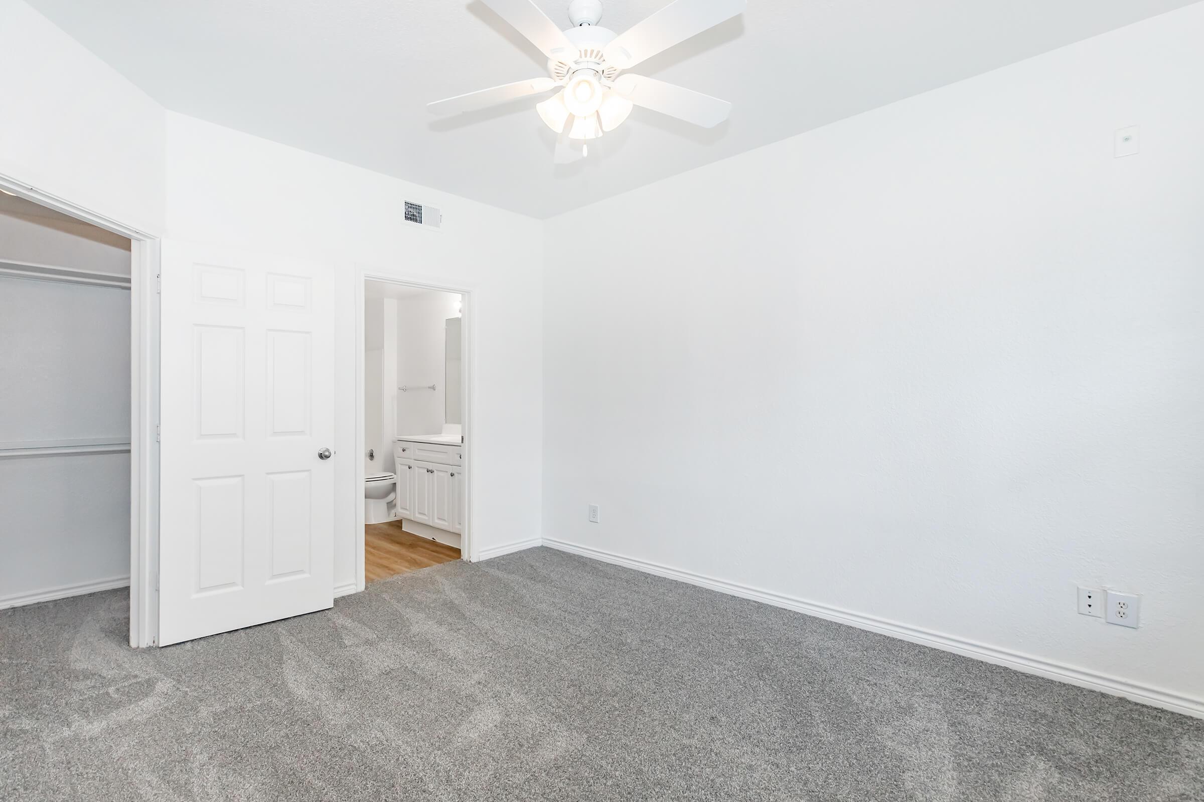 A clean, well-lit bedroom featuring light gray carpeting, a ceiling fan, and white walls. There is a closed closet with double doors on the left and a doorway leading to a bathroom in the background. The space is uncluttered, creating a serene atmosphere.