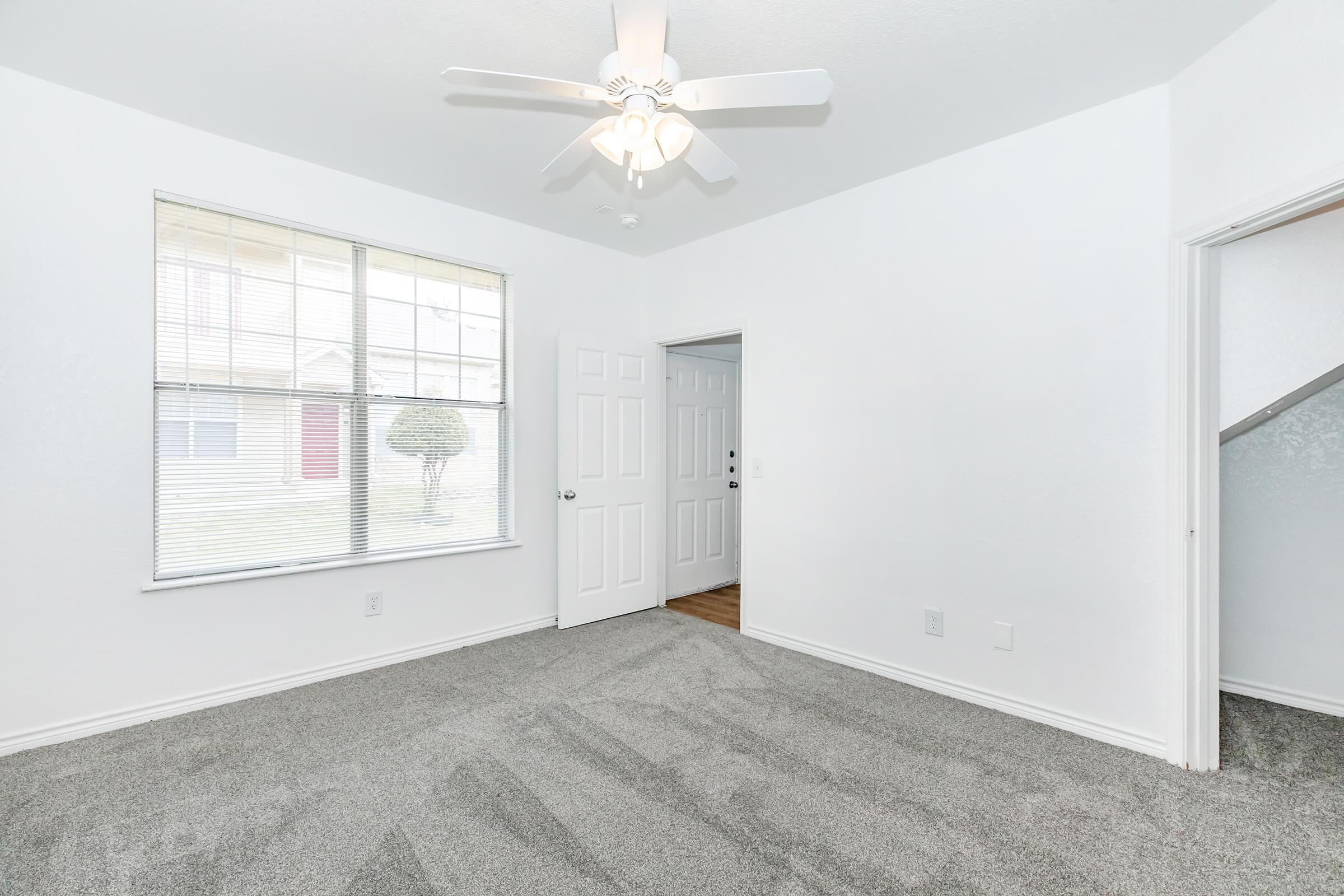 Bright, empty room with light gray carpet and white walls. A ceiling fan is installed, and there is a large window with blinds allowing natural light. The room features a door leading to another area and a view of the exterior with greenery outside. The overall atmosphere is clean and inviting.